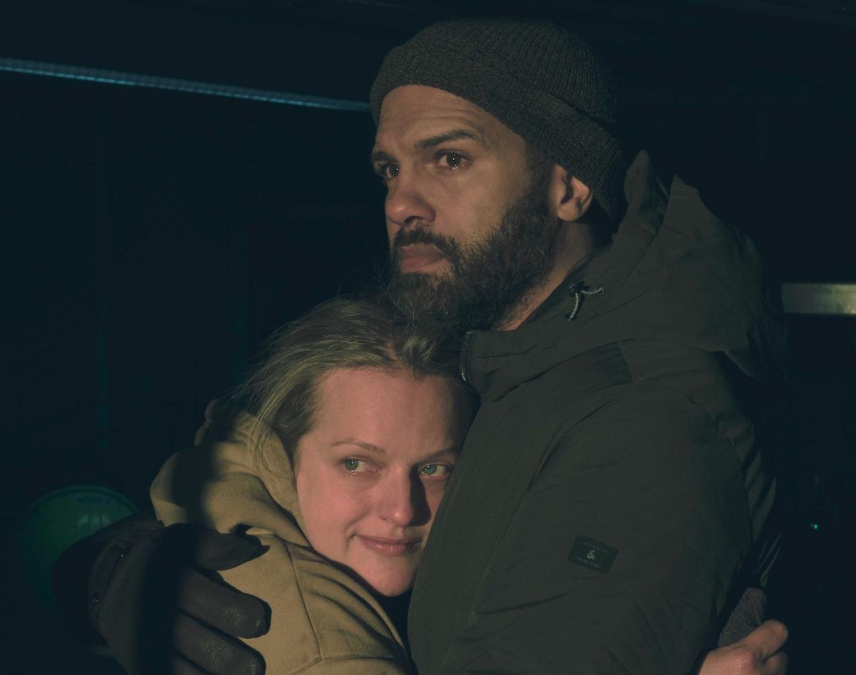 Elisabeth Moss and O-T Fagbenle as June and Luke in 'The Handmaid's Tale' Season 4 Episode 6. It's night and they're hugging on a boat, the background isn't seen because of the lighting. Moss wears a yellow hoodie, Fagbenle wears a green winter coat, gloves, and a beanie. Moss has a slight smile on her face and Fagbenle looks relieved.