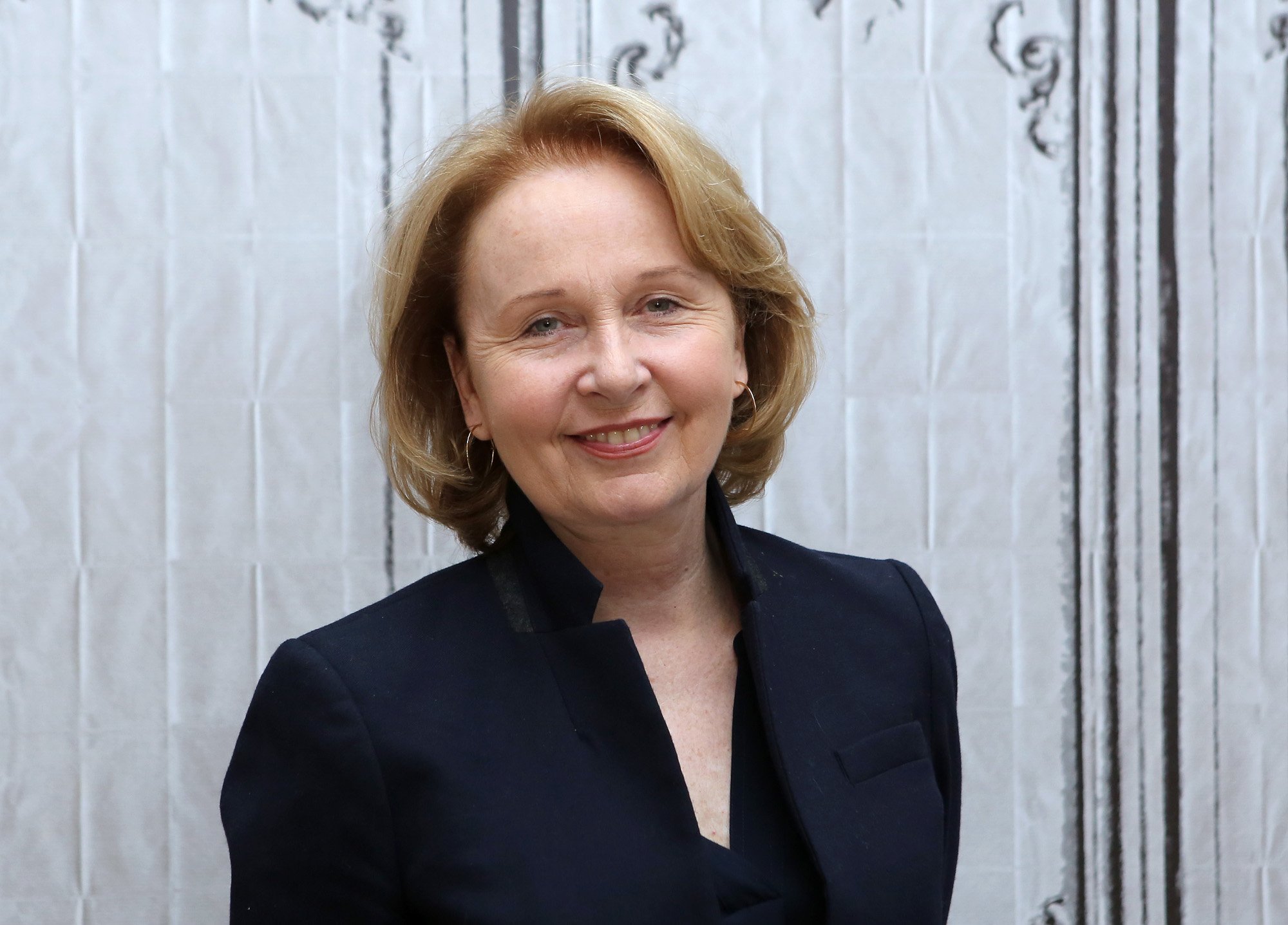 Kate Burton, who plays Meredith Grey's mother on Greys Anatomy, wearing a black blazer and smiling at the camera. Her hair is down and she's standing in front of a white wall.