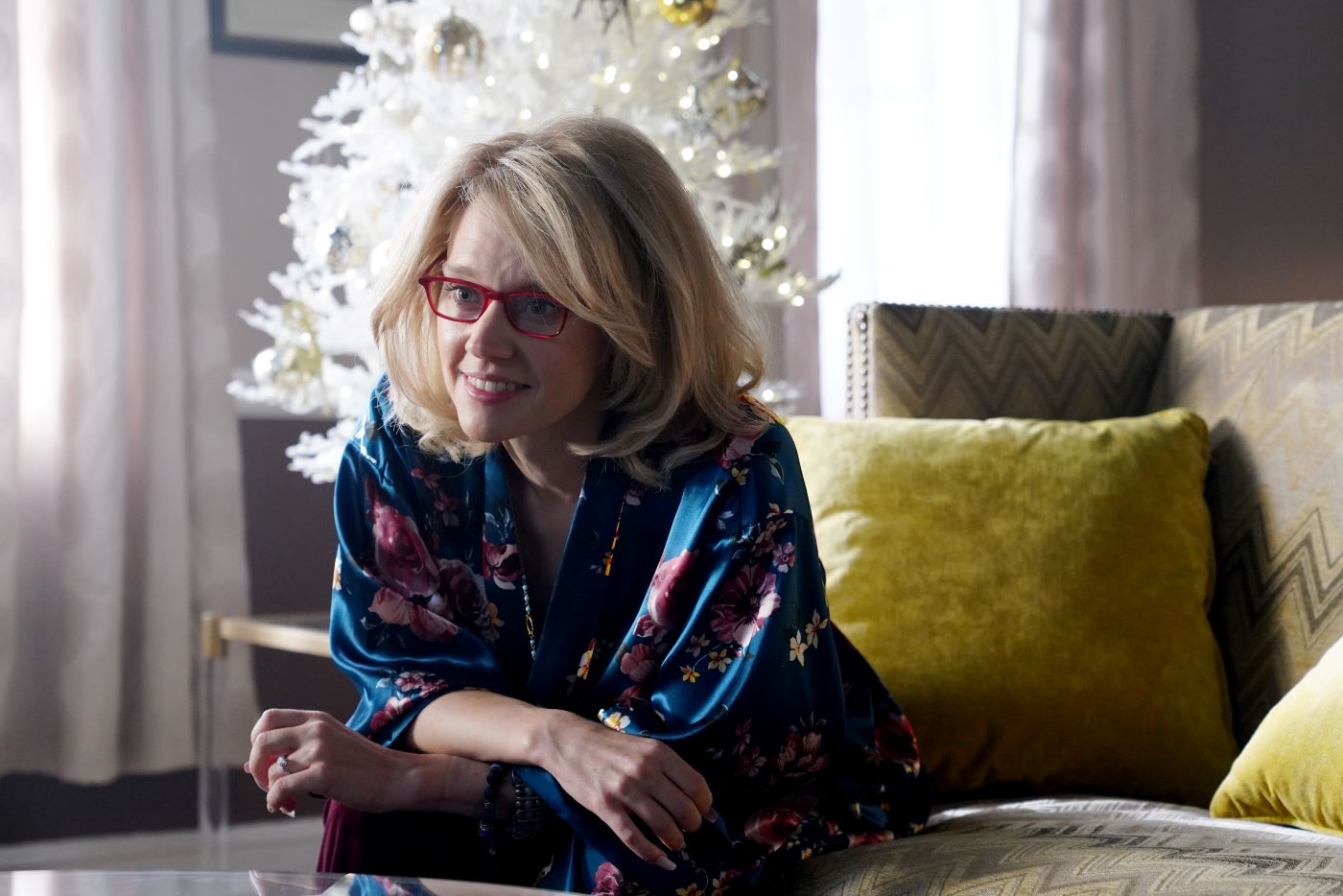 Kate McKinnon sitting on a green and grey coach with a white Christmas tree in the background wearing a blue shirt with a pink pattern on it.