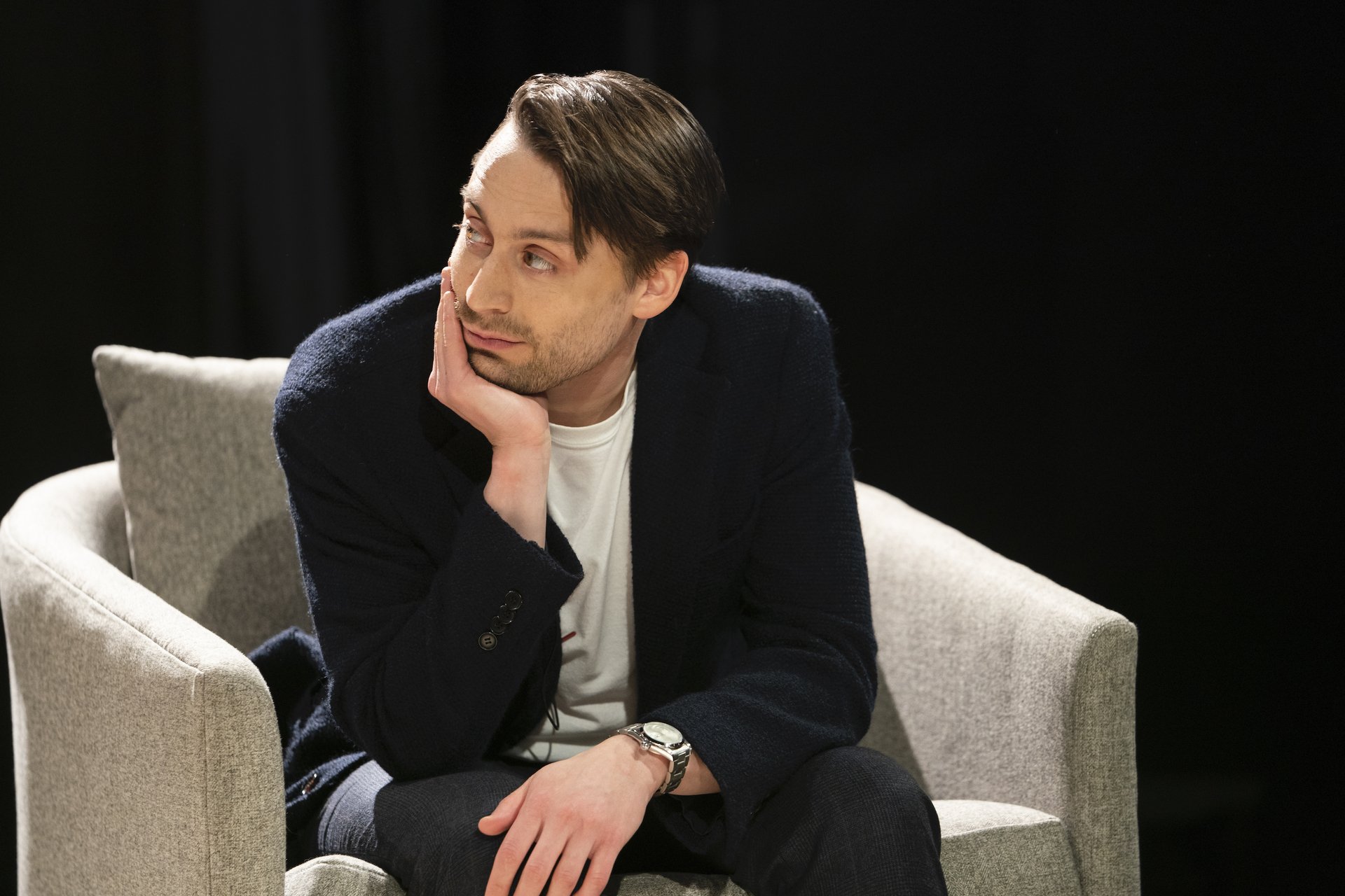 Kieran Culkin wears a black sweater and sits in a gray arm chair in 'Succession.'