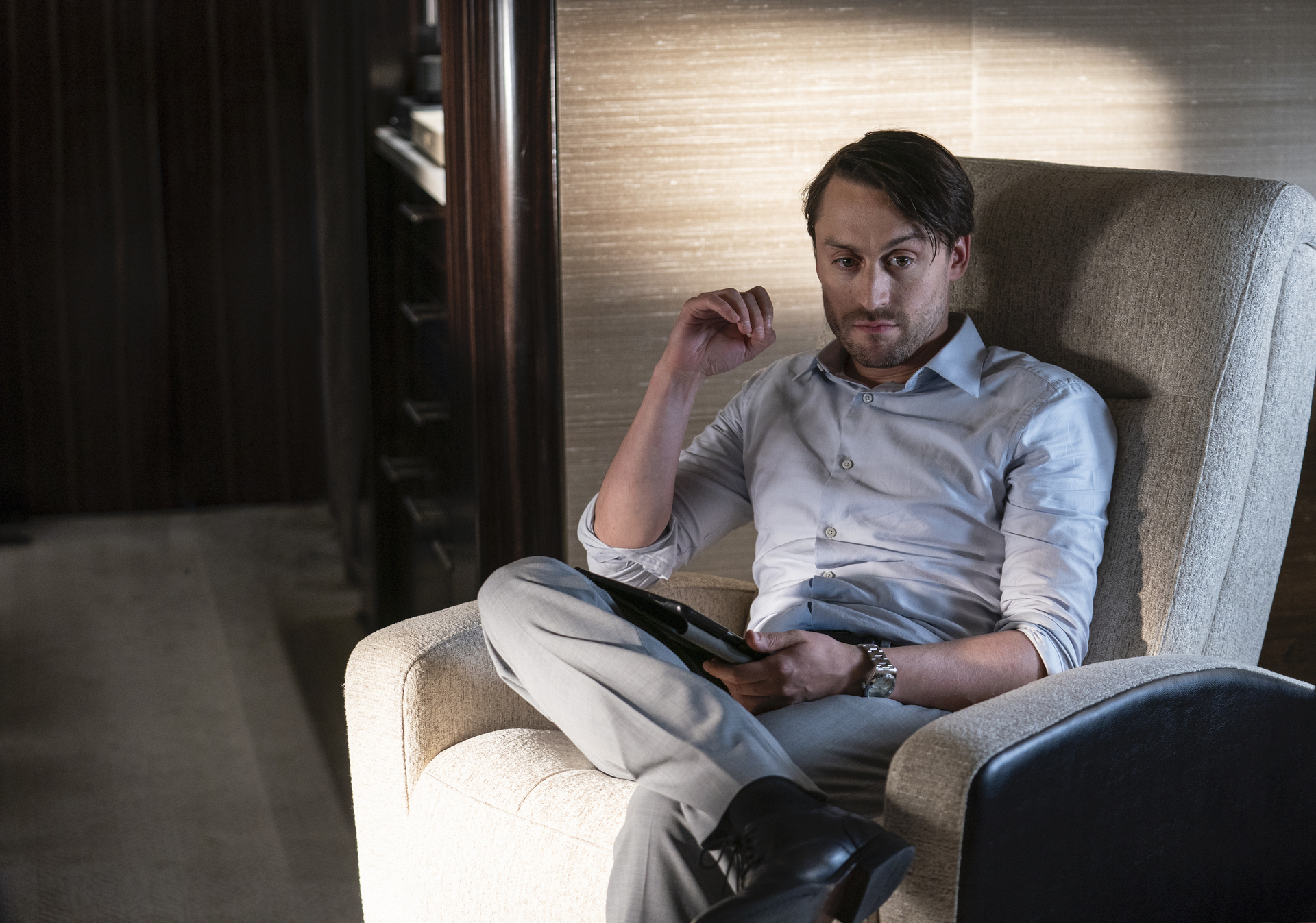 Kieran Culkin wears a gray shirt and sits in a gray chair, holding an iPad in 'Succession.' 