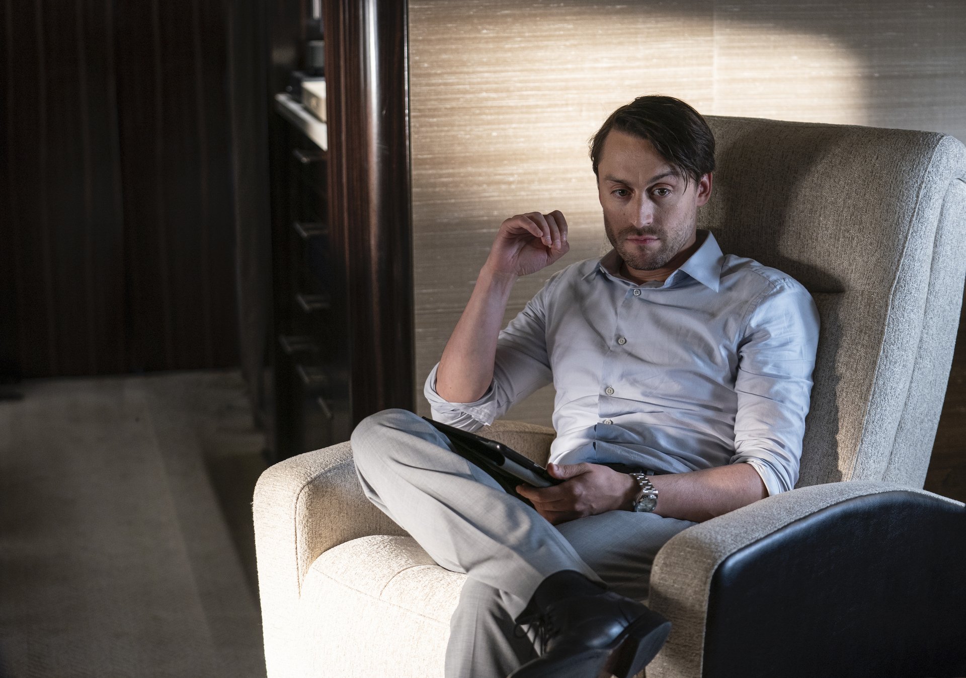 Kieran Culkin wears a gray shirt and sits in a gray chair, holding an iPad in 'Succession.'