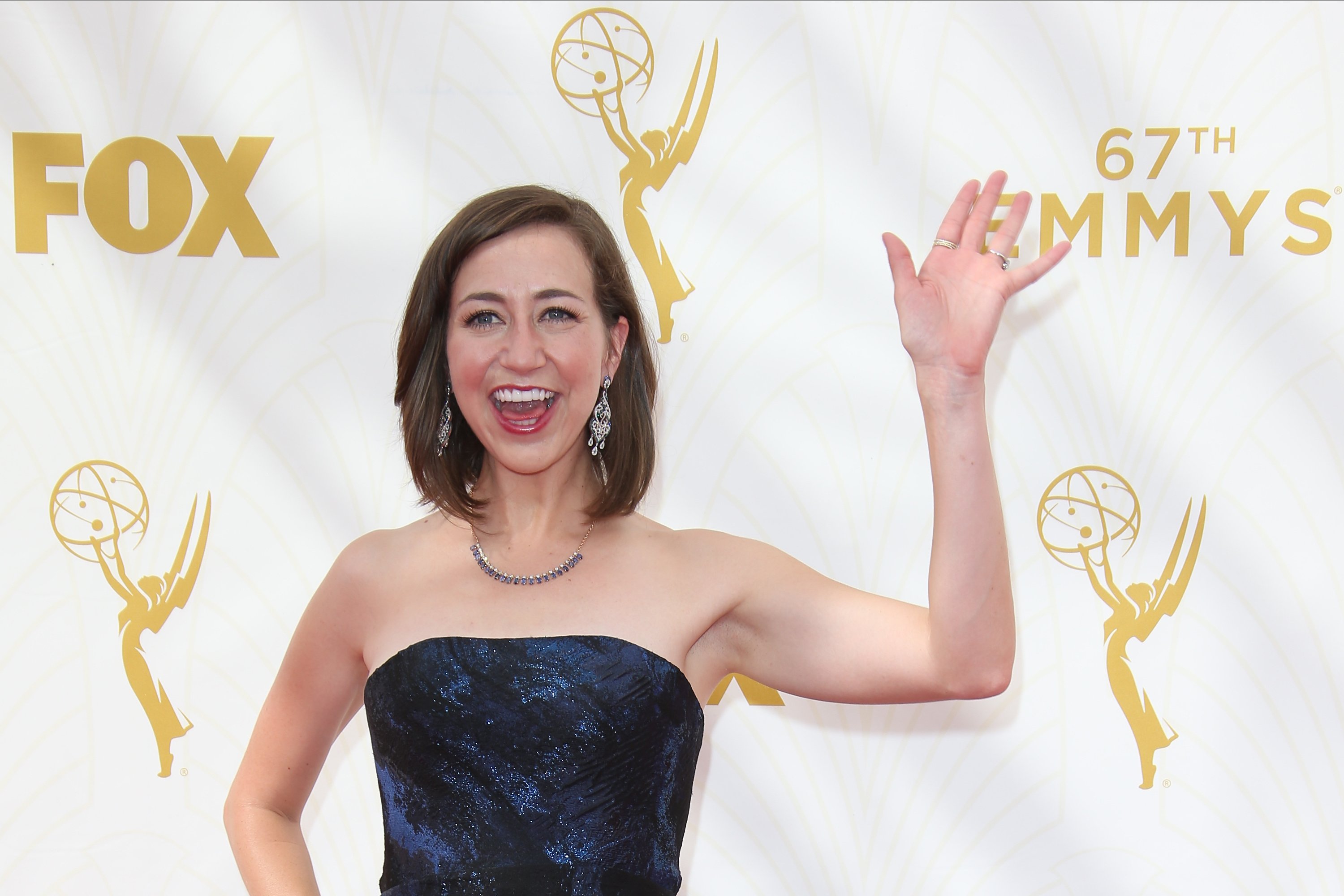 Kristen Schaal waves wearing a blue strapless dress and earrings.