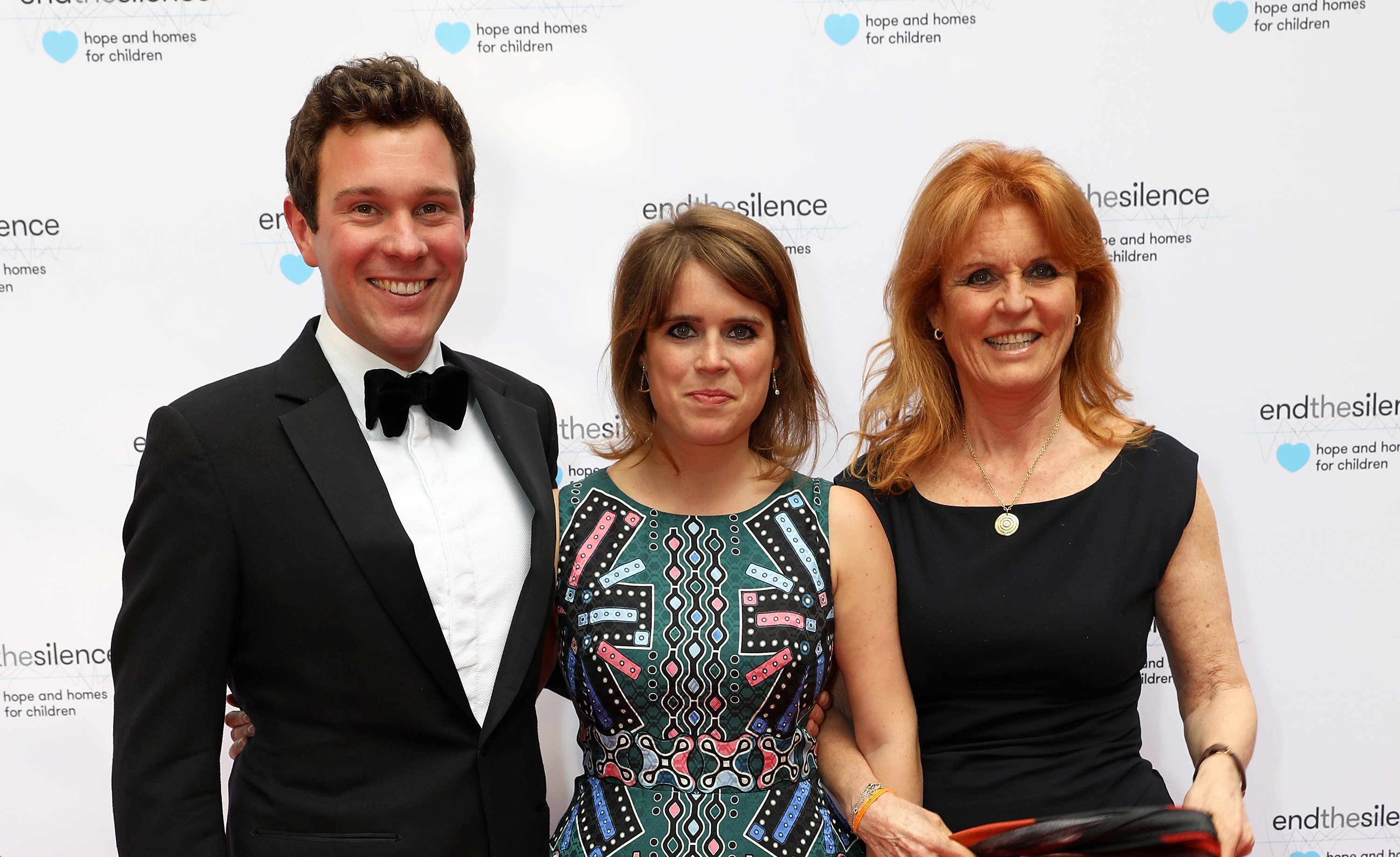 (L to R): Jack Brooksbank, Princess Eugenie, and Sarah Ferguson posing together on the red carpet at an event