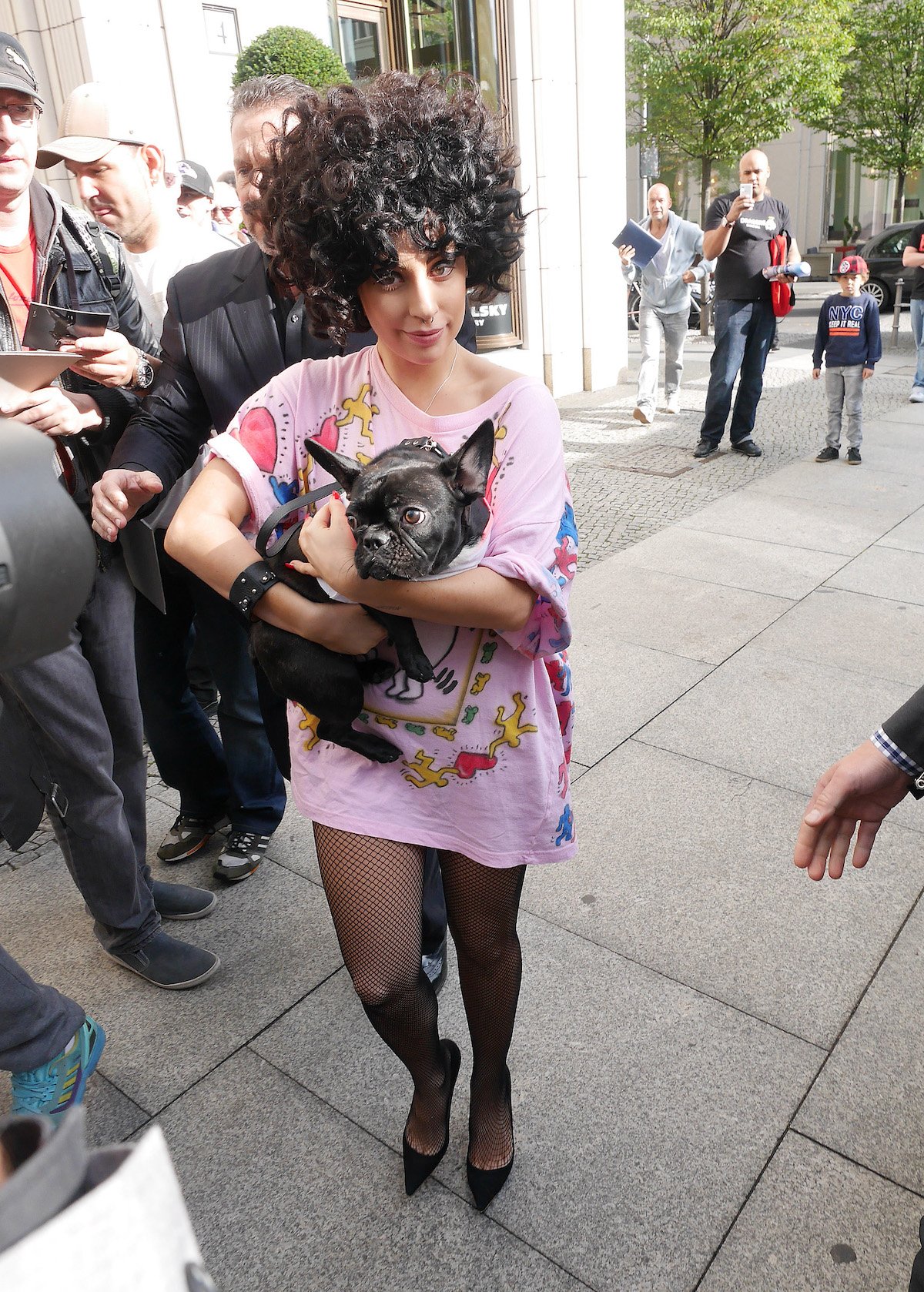 Lady Gaga in an oversized pink shirt and curly black wig holding a black dog.