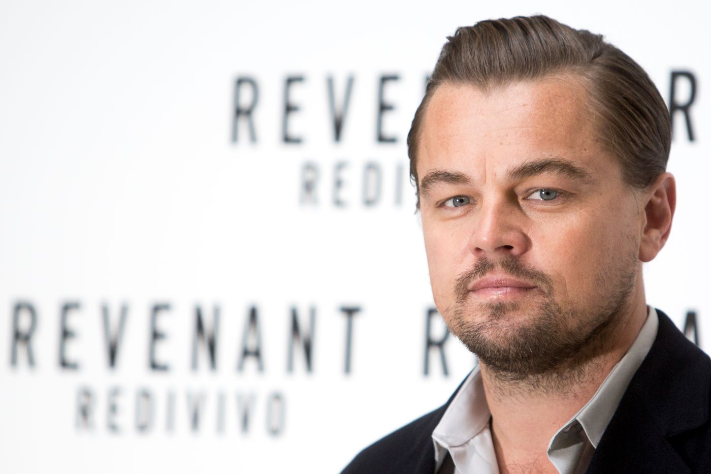 Leonardo DiCaprio, star of the revenant, dressed in a suit jacket and a collared shirt standing in front of a white background with Revenant written in black text.