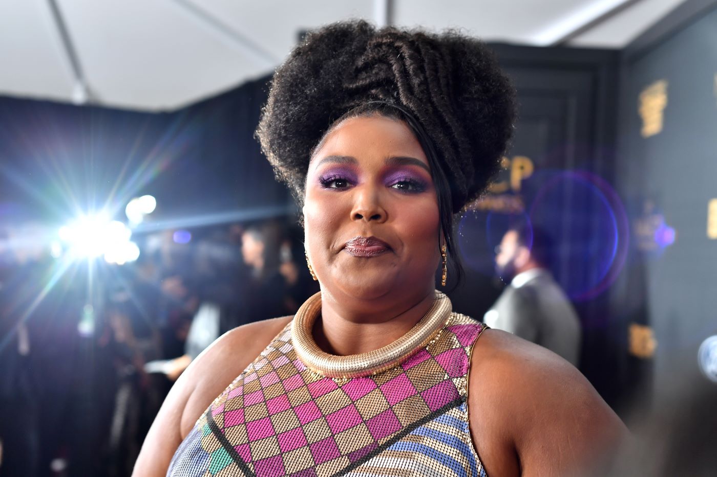Lizzo is wearing a colorfully patterned top standing in front of a black background with gold writing and a bright light on the left side.