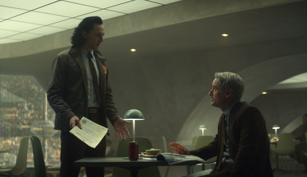 Tom Hiddleston and Owen Wilson in 'Loki' Season 1. Hiddleston stands at the table Wilson is sitting at. The walls are beige with curved archways. Hiddleston and Wilson appear to be disagreeing.