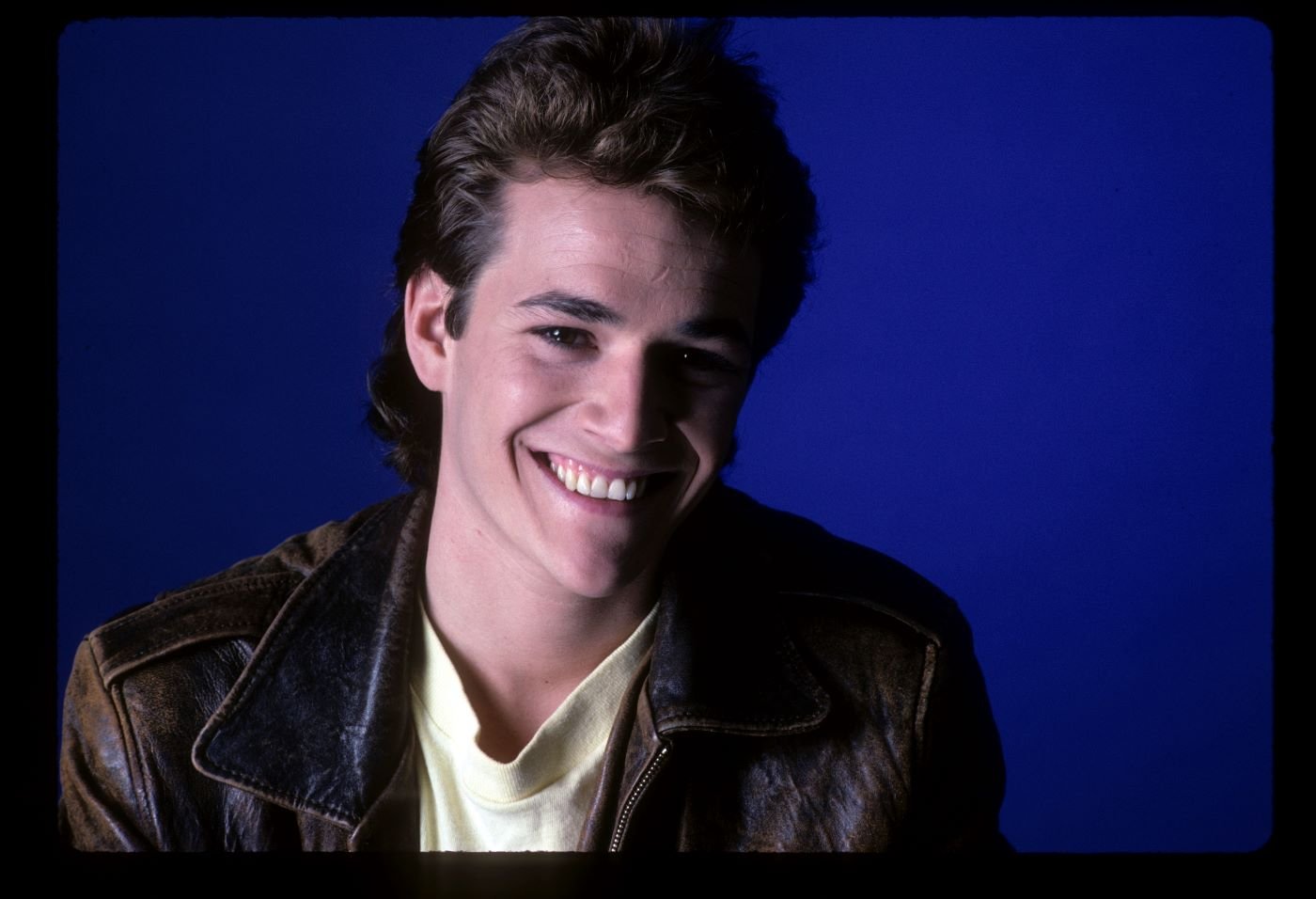 Luke Perry in a black leather jacket with a cream undershirt in front of a solid blue background.