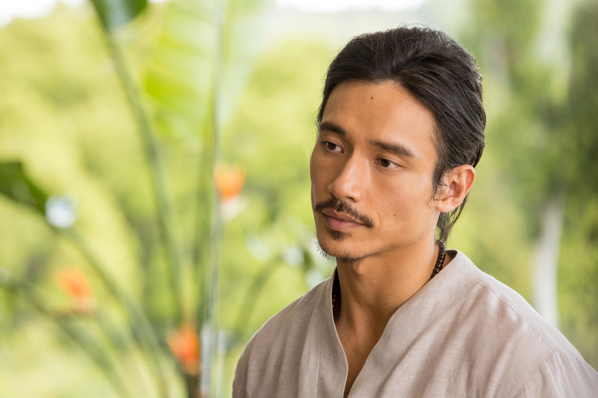 Manny Jacinto as Yao in 'Nine Perfect Strangers.' His hair is pulled back into a ponytail and he's wearing a beige shirt. His head is tilted and he's standing in front of greenery.