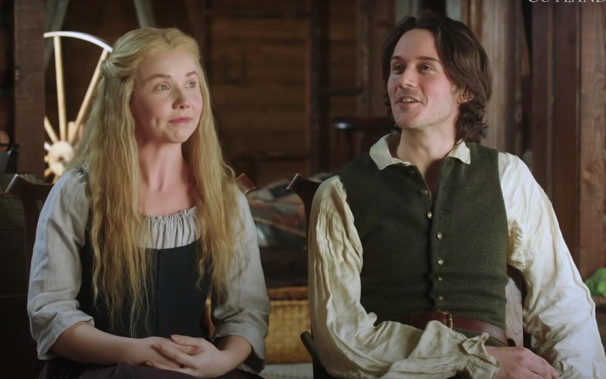 Lauren Lyle and César Domboy dressed as 'Outlander's Marsali and Fergus in a promotional video for 'Outlander' Season 6. Lauren sits on the left in a green colonial dress with a beige quarter-sleeve top underneath. Her long blonde hair is down in front of her shoulders. Domboy sits on the right wearing a tan colonial shirt and green vest with gold buttons and a brown leather belt. They sit inside in a log cabin. Marsali and Fergus' newest son, Henri Christian, could have a big plot in the upcoming 'Outlander' Season 6.