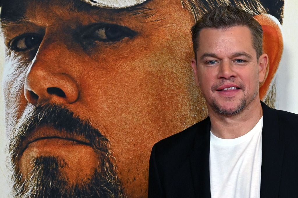 Matt Damon poses in front of a 'Stillwater' photo. He's wearing a black suit and white shirt.
