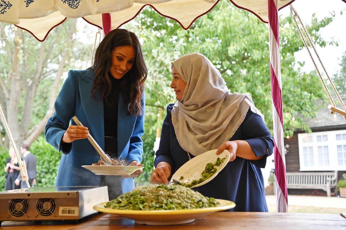 Meghan Markle in a blue blazer plating food with tongs while speaking with a woman