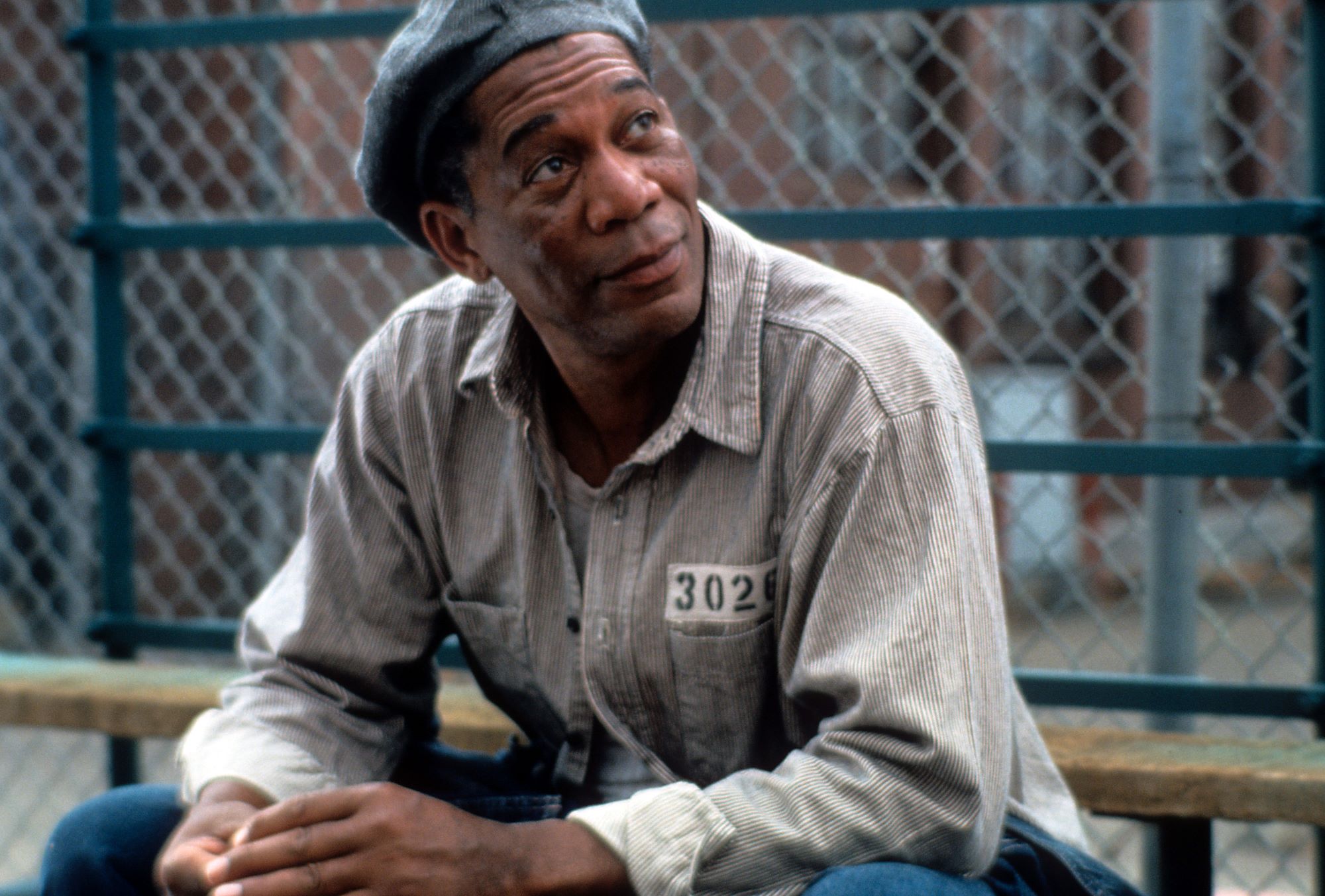 Morgan Freeman in a scene from 'The Shawshank Redemption' sitting in his prison uniform and hat on top of a picnic table outside in the yard.