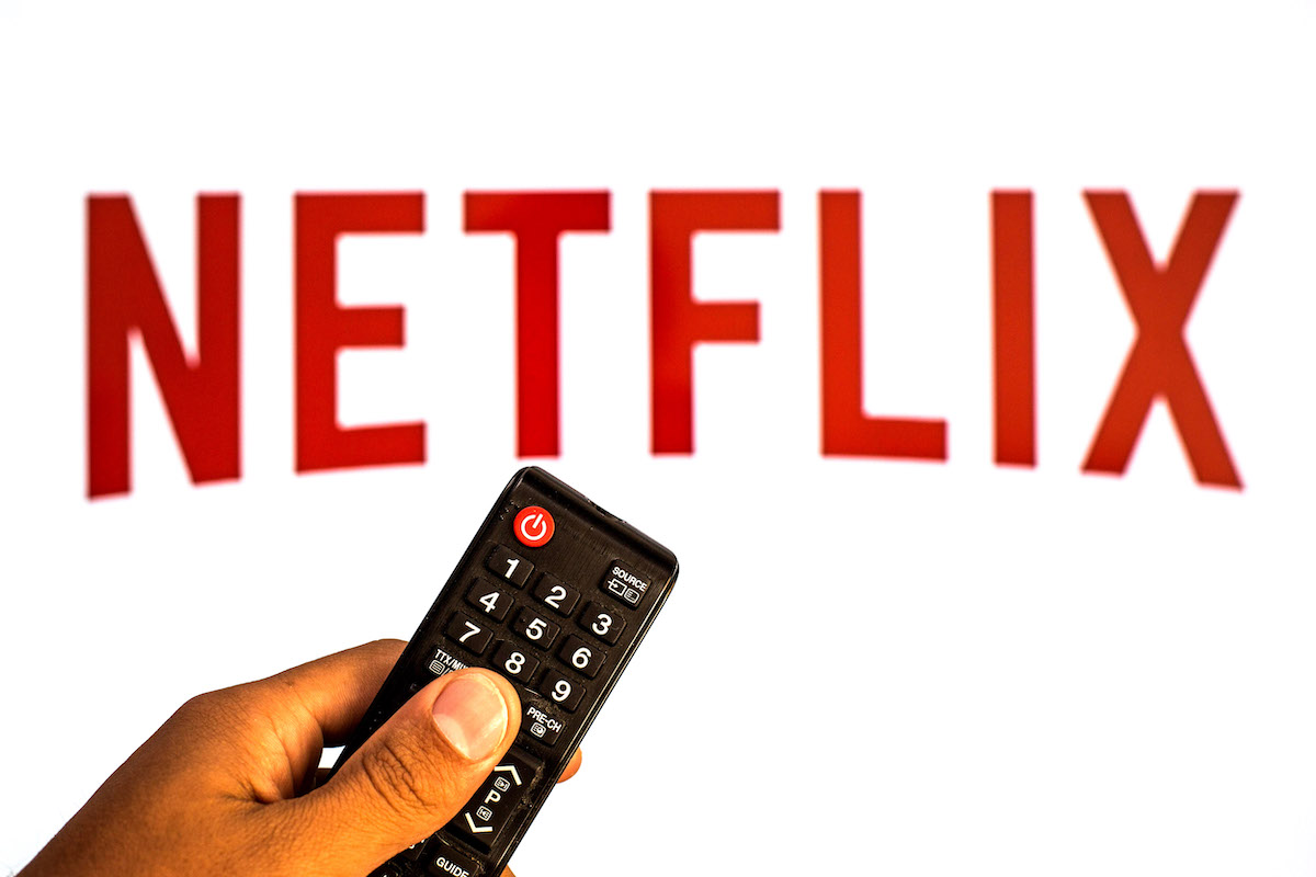 The Netflix logo in red letters in front of a white background with a hand holding a remote