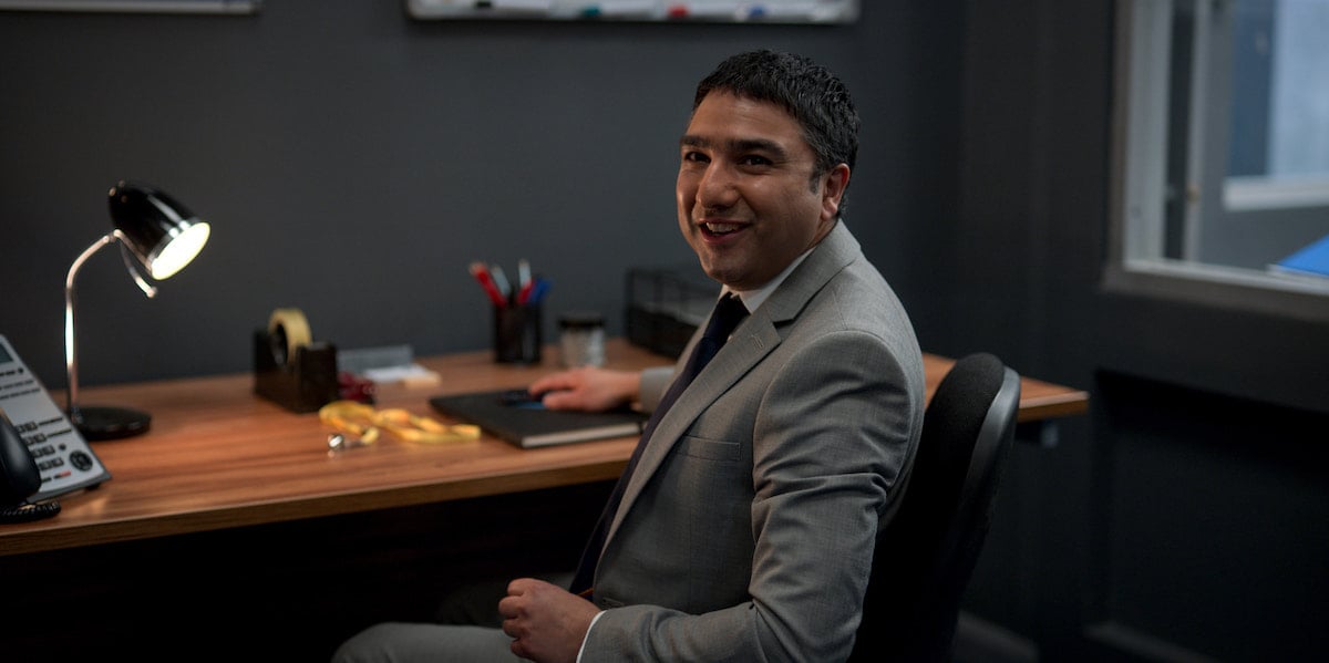 Nick Mohammed smiles as he sits at a desk wearing a suit and tie as Nathan Shelley in 'Ted Lasso' Season 2