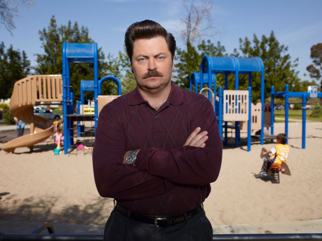 Nick Offerman as Ron Swanson stands in front of the park wearing a burgundy sweater with his arms crossed and a stern look on his face.
