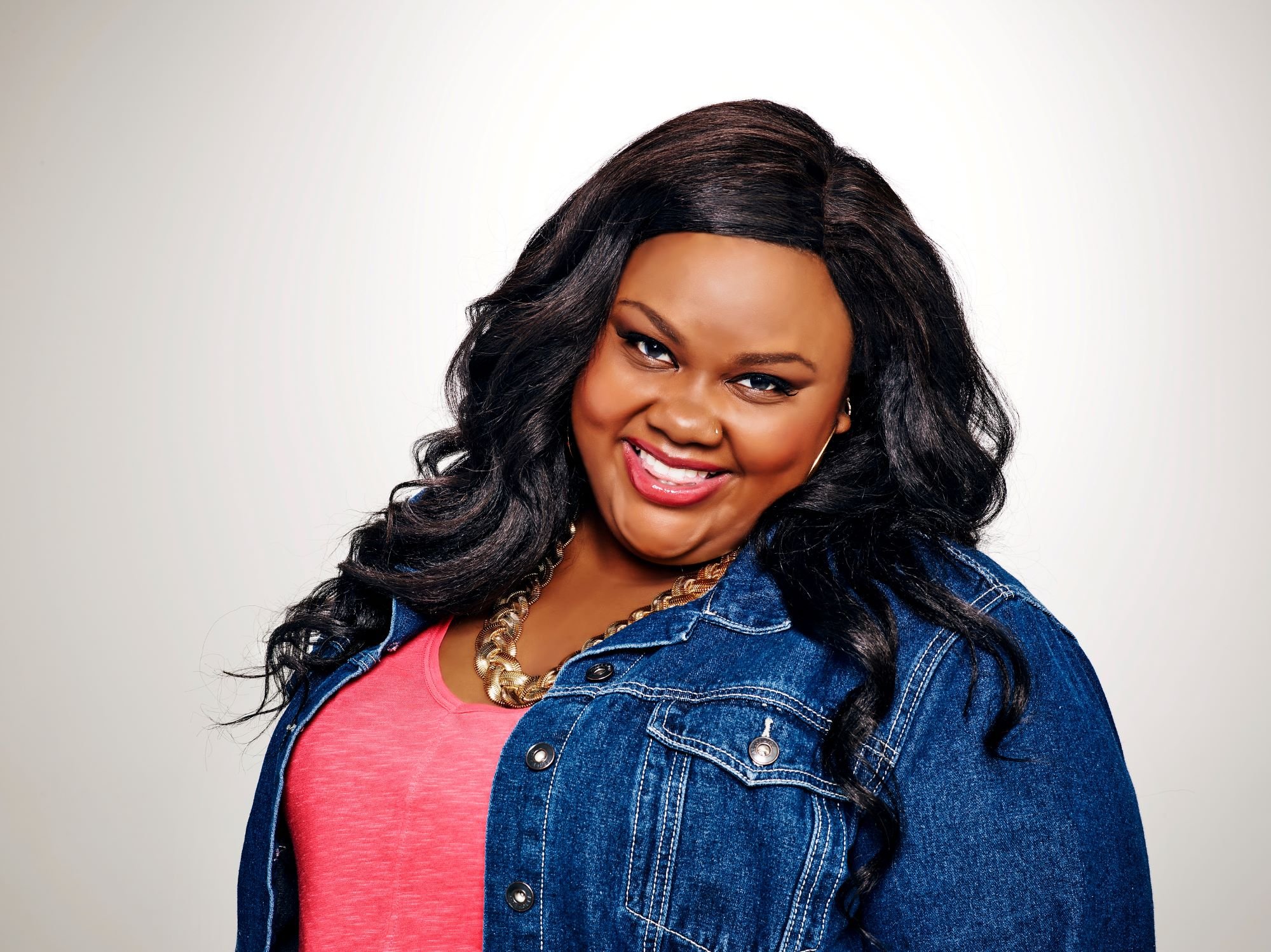 Nicole Byer dressed in a blue jean jacket in front of a white background.