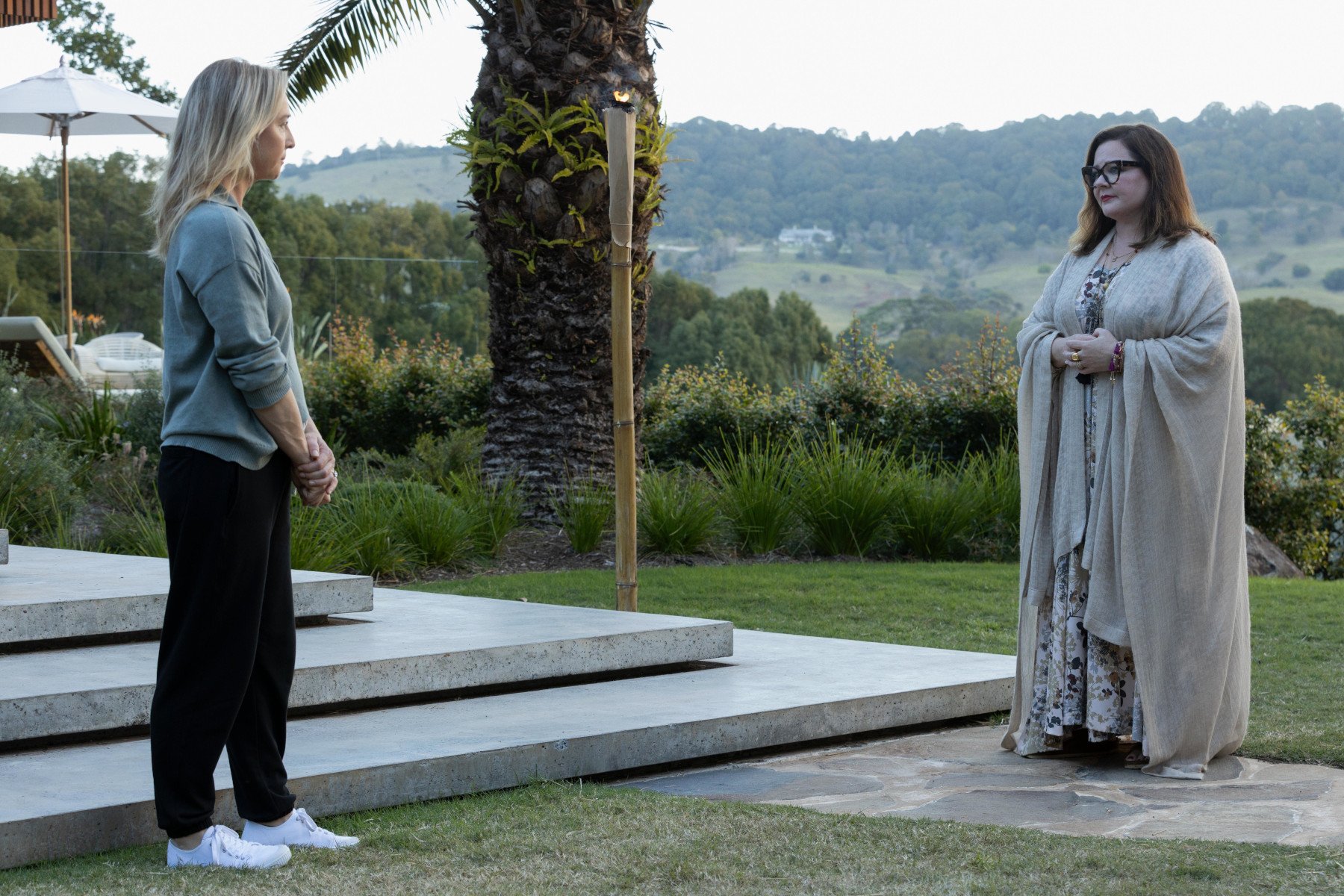 Heather (Asher Keddie) and Frances (Melissa McCarthy) standing across from one another in Hulu's 'Nine Perfect Strangers' Episode 1. The two have their hands clasped in front of each other and look uncomfortable. There are trees and greenery in the background.