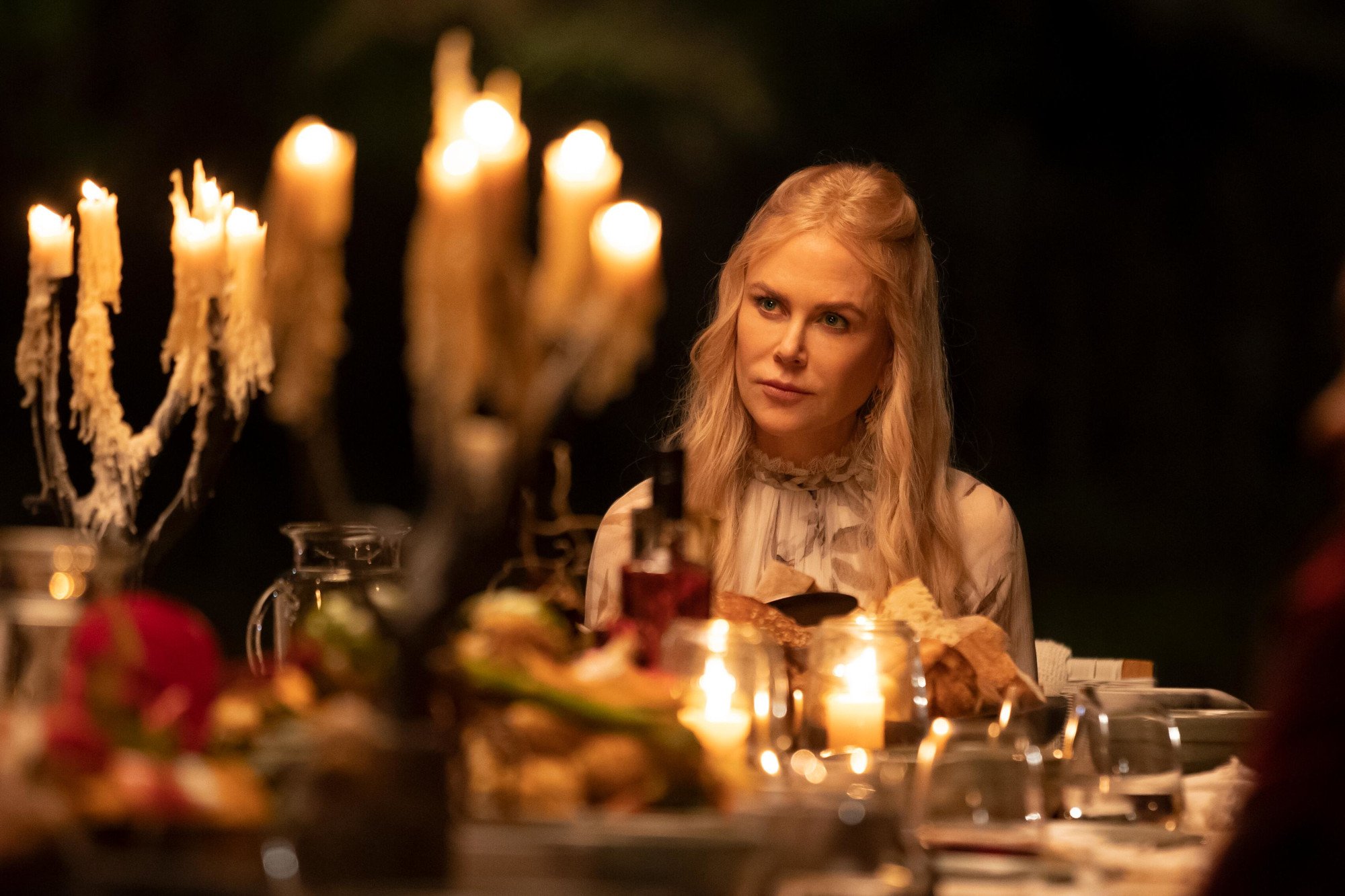 Nicole Kidman as Masha in 'Nine Perfect Strangers' Episode 4. She's sitting at the edge of a dinner table with candles on it. Her head is tilted like she's listening to someone talking.