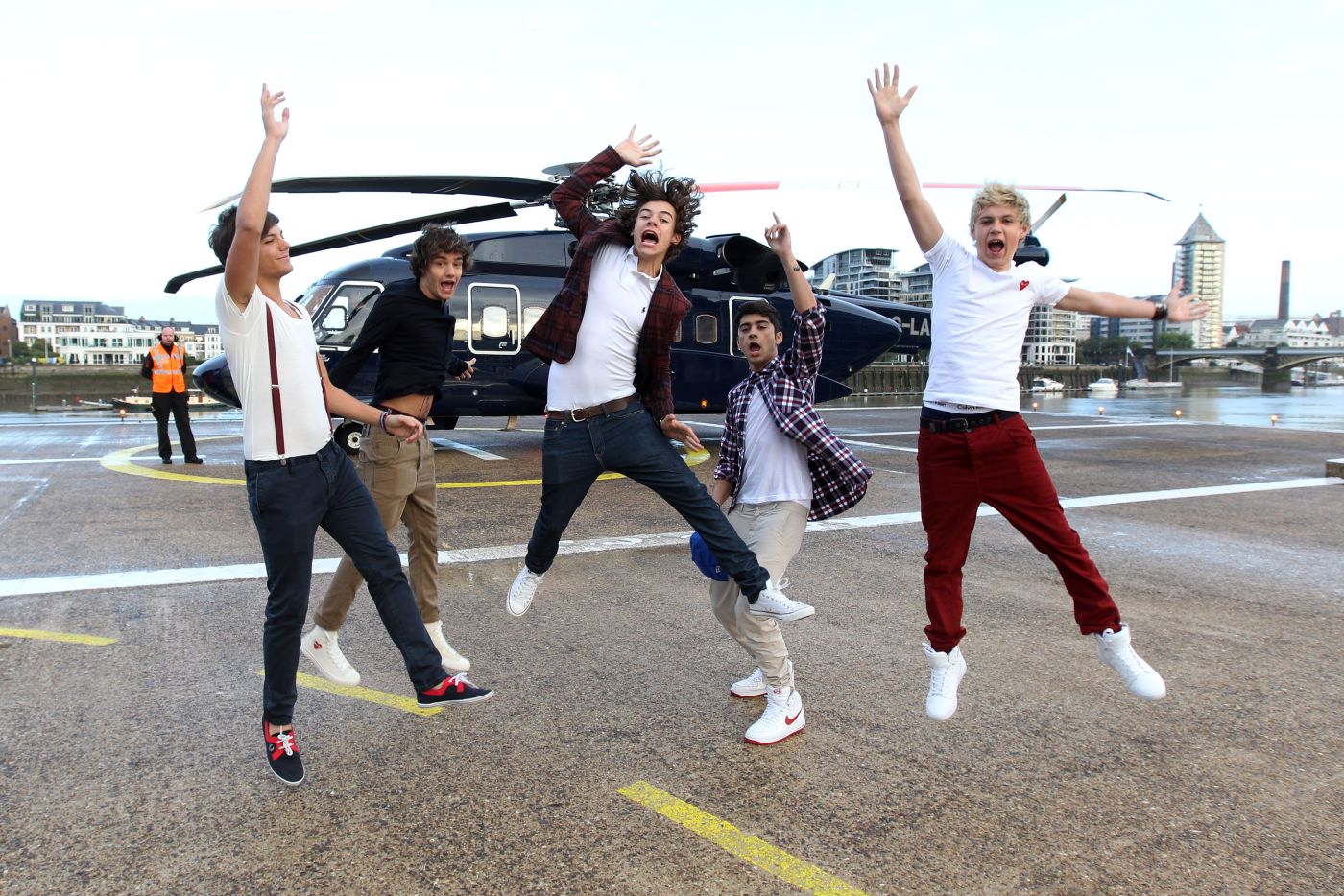 All five One Direction band members jumping in the air in front of a helicopter in various poses.