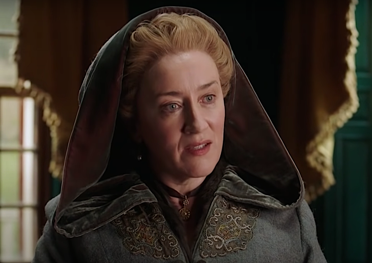 'Outlander's Maria Doyle Kennedy as Aunt Jocasta in a World 'Outlander' Day video. She wears a blue/grey colonial cloak with her hood up, a brown ribbon with a pendant around her neck. She wears a blonde wig styled in a traditional women's colonial style, and she sits in front of a window with a draping yellow curtain. Kennedy will likely return in the 'Outlander' Season 6 cast, but she could potentially not come back for season 7, which has already been green-lit by Starz.