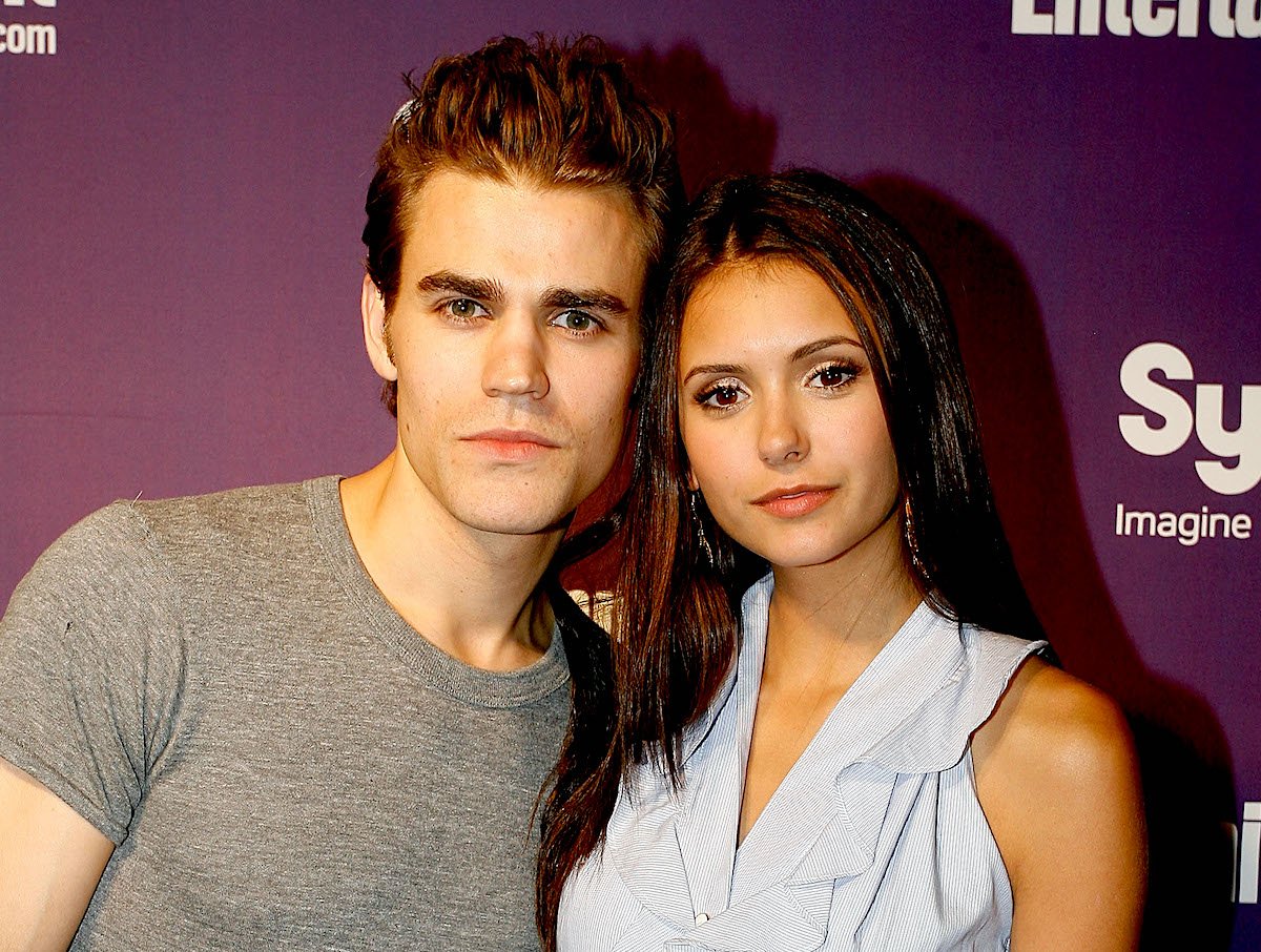 'The Vampire Diaries' stars Paul Wesley and Nina Dobrev stand closely together as they pose in front of a purple background. Wesley wears a grey T-shirt, Dobrev wears a sleeveless, collared dress that's baby blue on top and black on bottom.