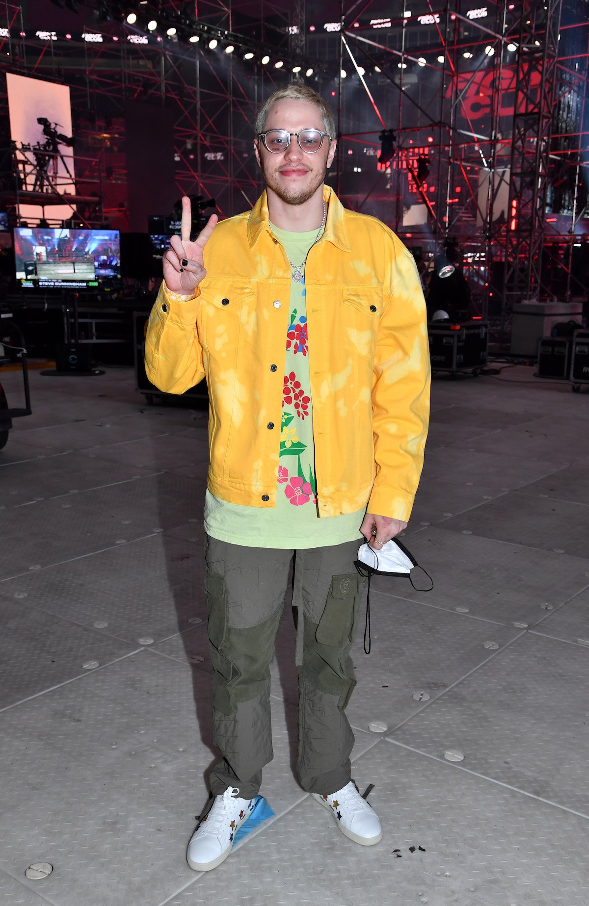 Pete Davidson, dressed casually in a yellow jacket, flashes a peace sign at the camera and smiles.