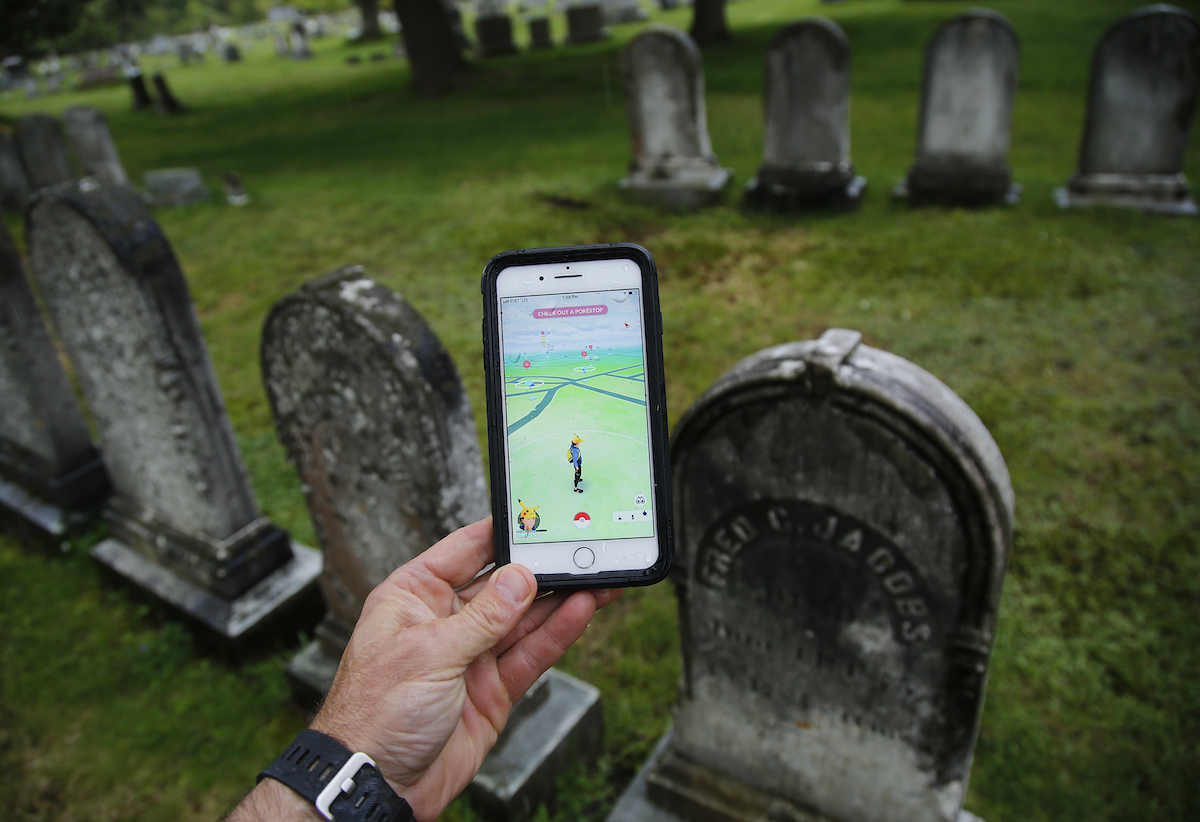 A hand holds a phone playing Pokemon Go. The background clearly shows the player is standing in a graveyard before a row of tombstones.