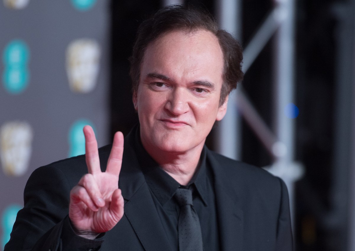 Quentin Tarantino wears a black suit and holds his hand up in a peace sign on the red carpet