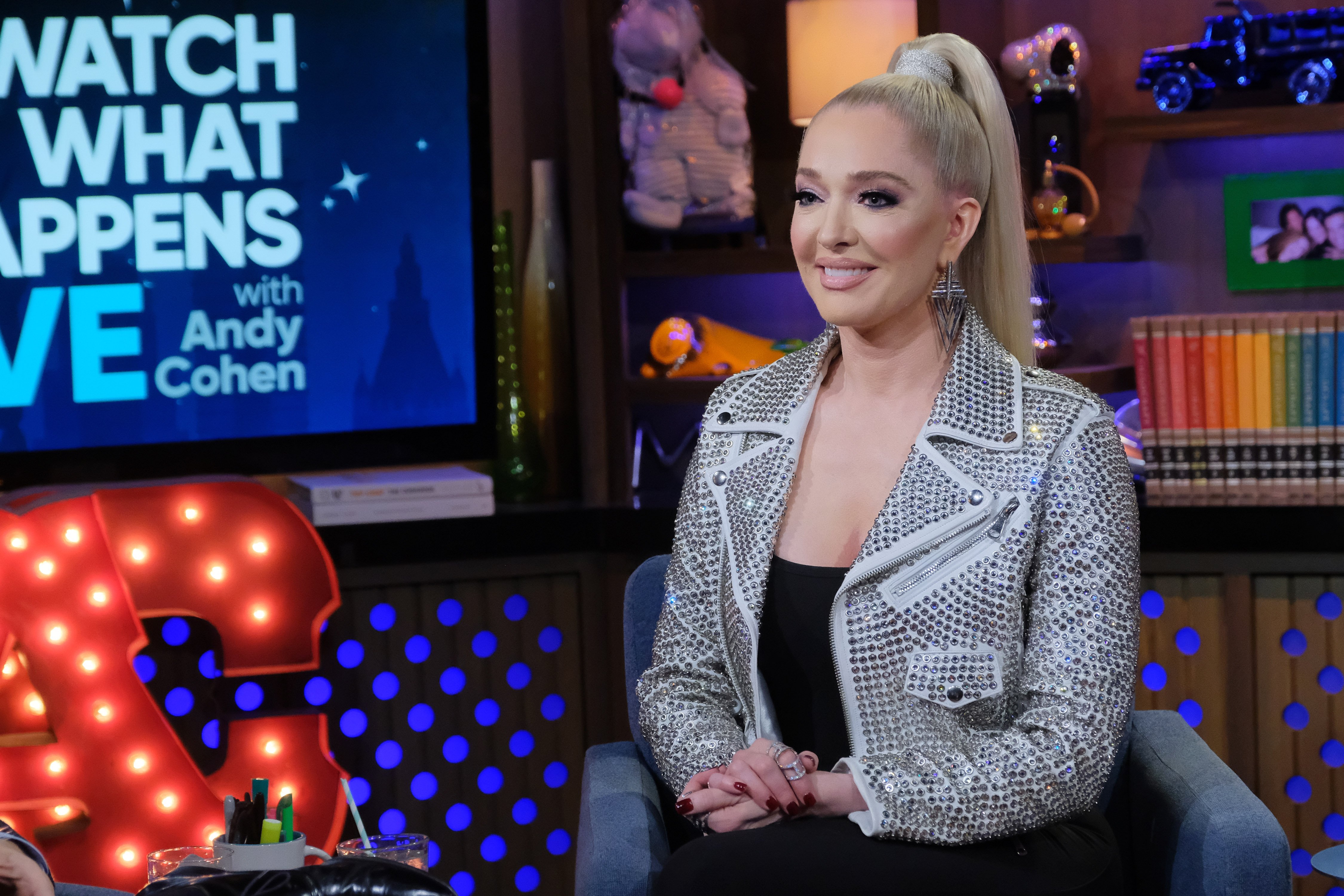 Erika Jayne smiling while wearing a silver jacket on the set of 'Watch What Happens Live'