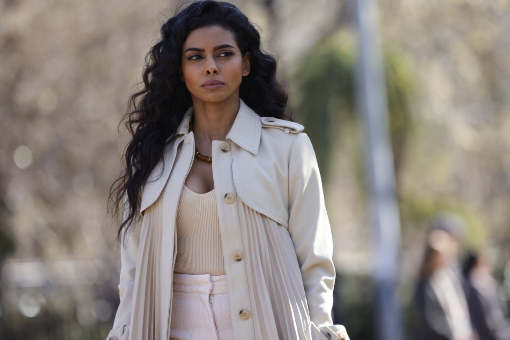 Rana Roy as Priya Laghari has a concerning look on her face as she walks through the park wearing all beige and white.