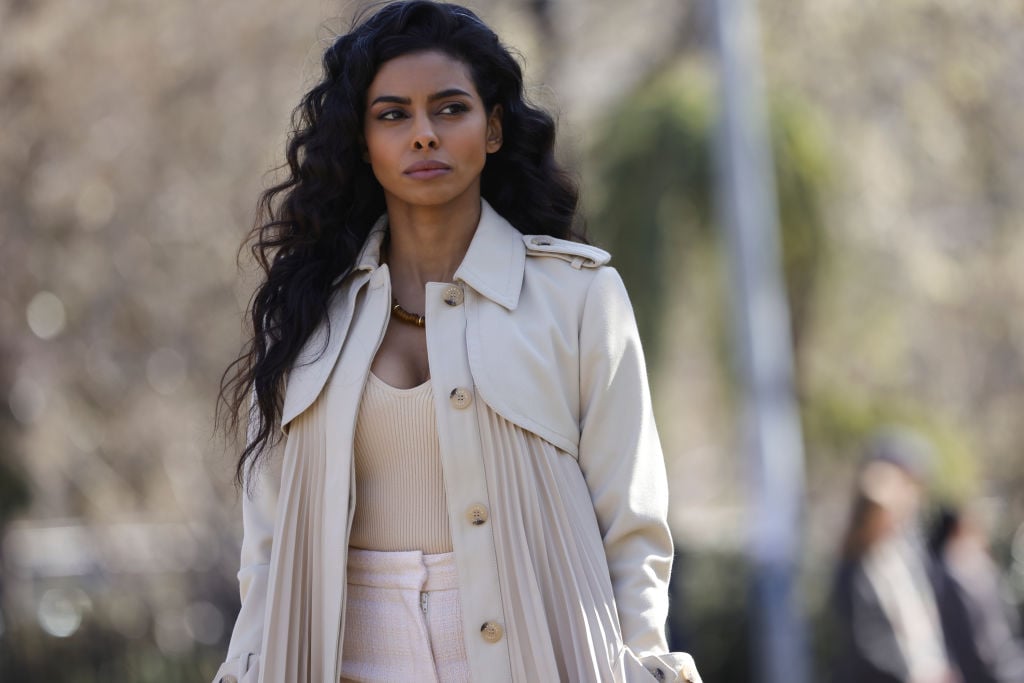 Rana Roy as Priya Laghari has a concerning look on her face as she walks through the park wearing all beige and white. 