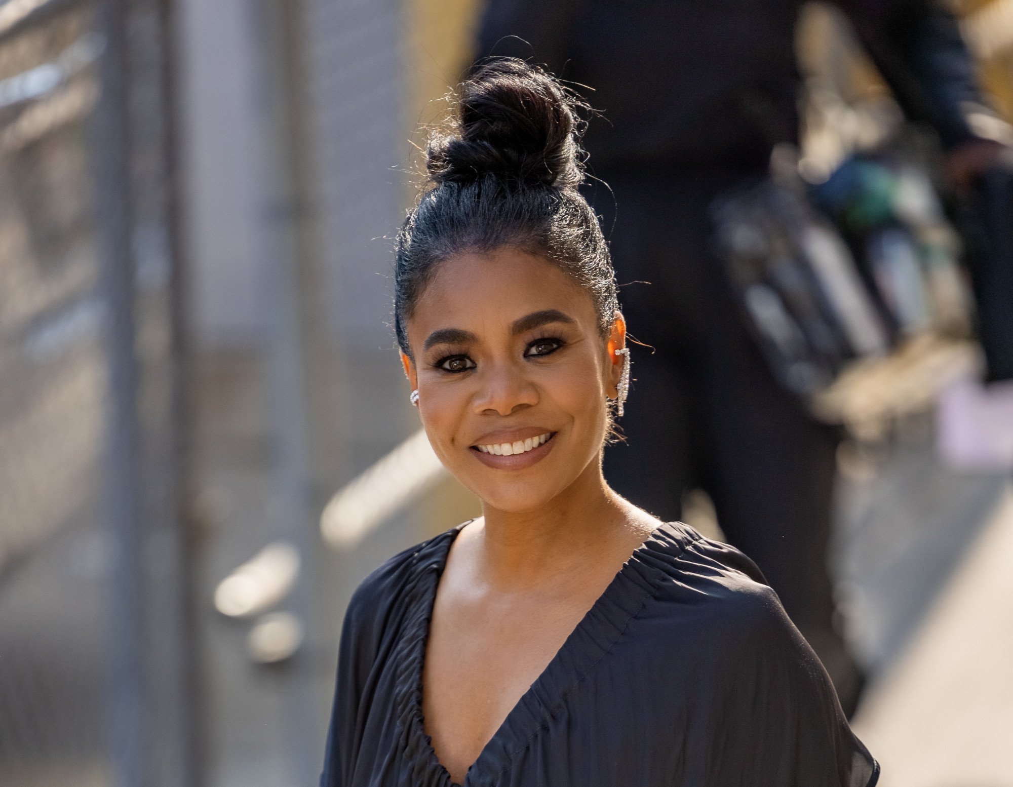 'Nine Perfect Strangers' cast member Regina Hall. She's walking outside and wearing a black outfit with a V-shaped neckline. Her hair is in a bun and she's smiling at the camera. .