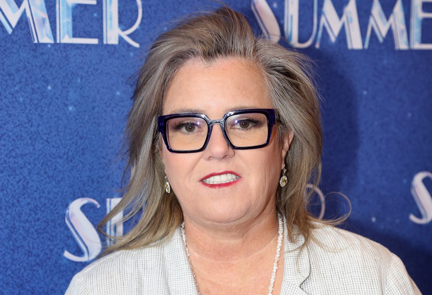 Rosie O'Donnell wearing a light beige colored jacket with a white undershirt against a blue and background with white writing on it.