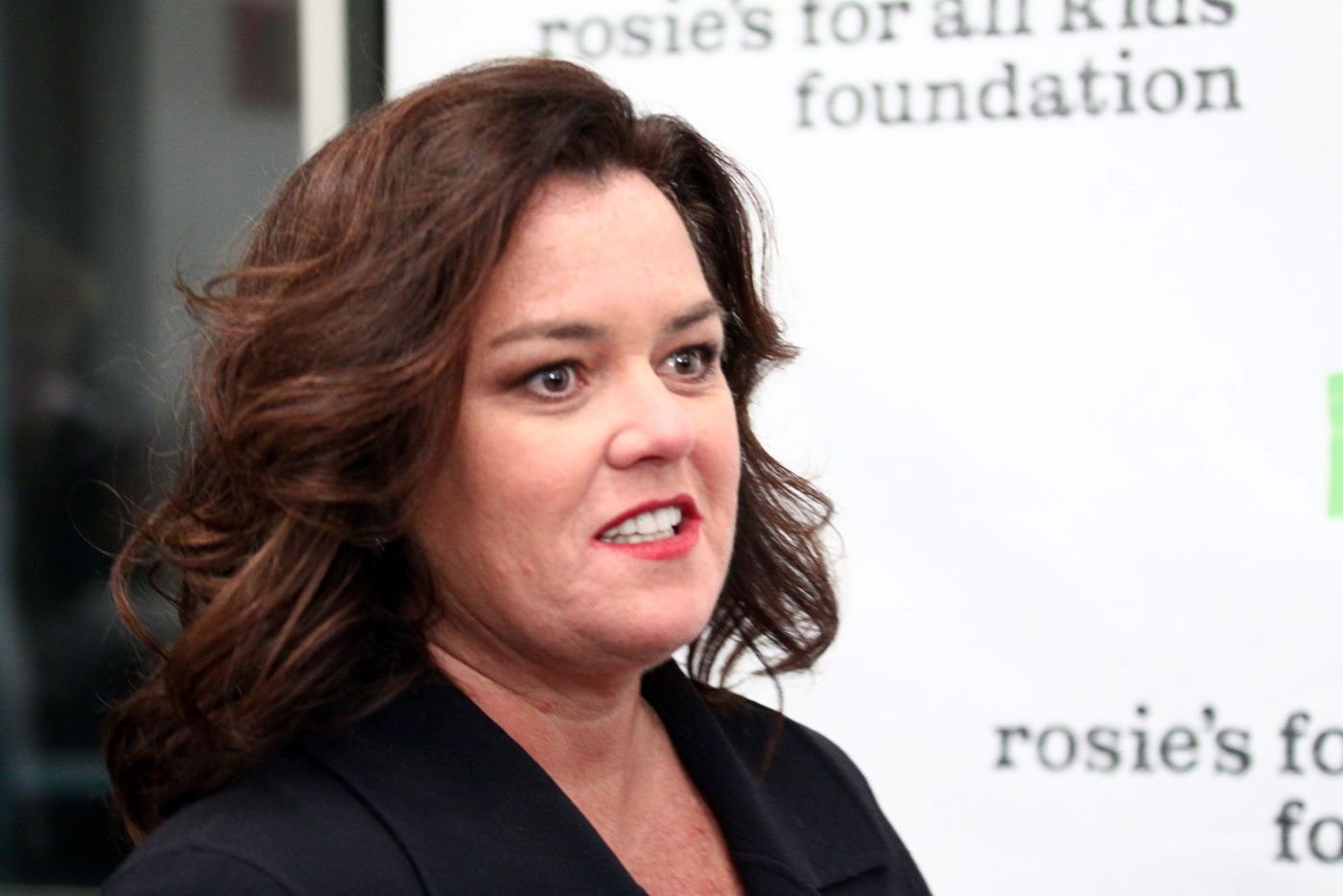 Rosie O'Donnell wearing a black jacket with a black undershirt in front of a white backdrop with black writing.
