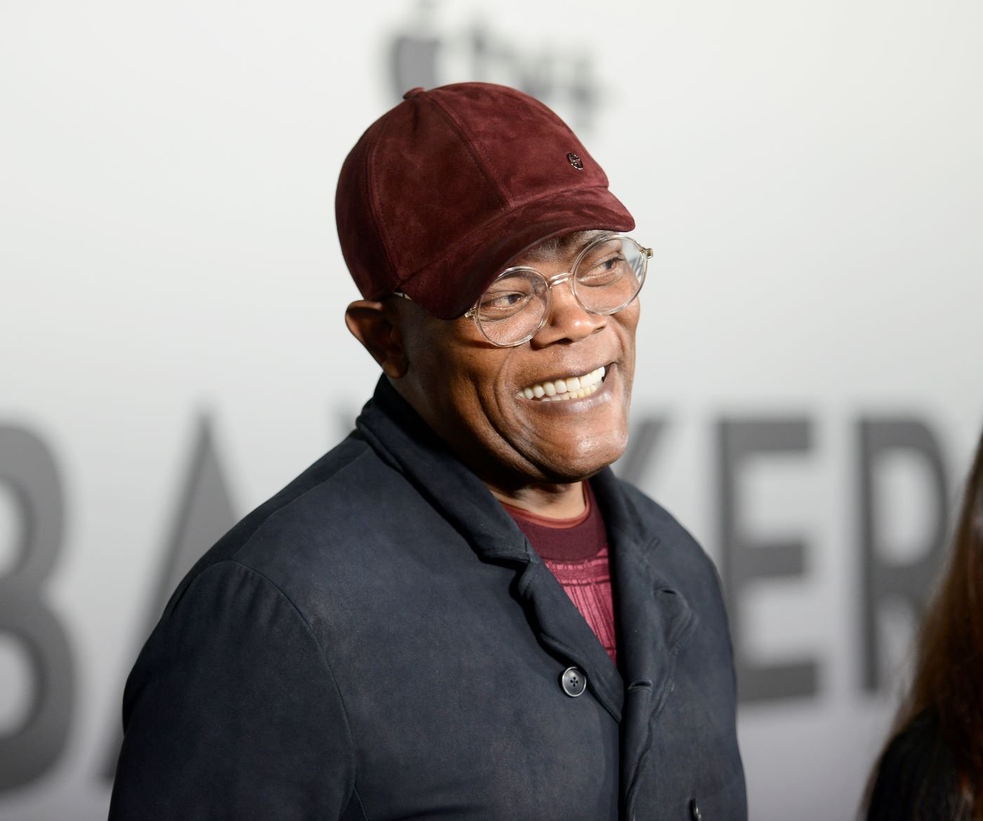 Samuel L. Jackson standing in front of a white and black background wearing a burgundy baseball hate and shirt with a black suit jacket.