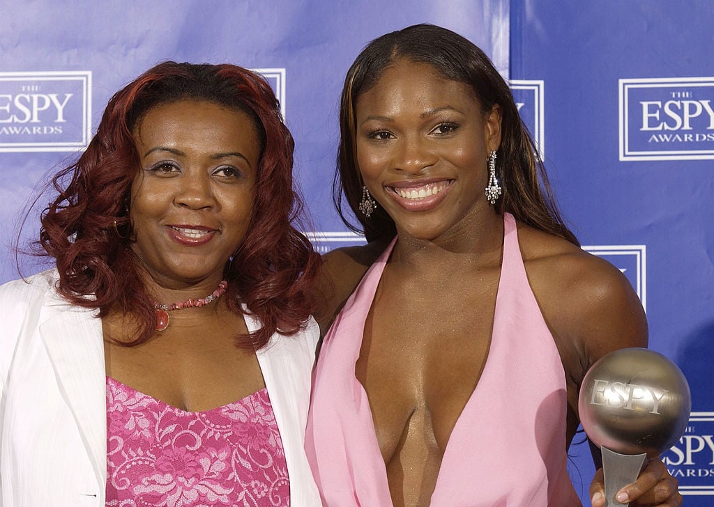 Serena Williams and Yetunde Price pose on stage all smiles. Both are wearing pink dresses.