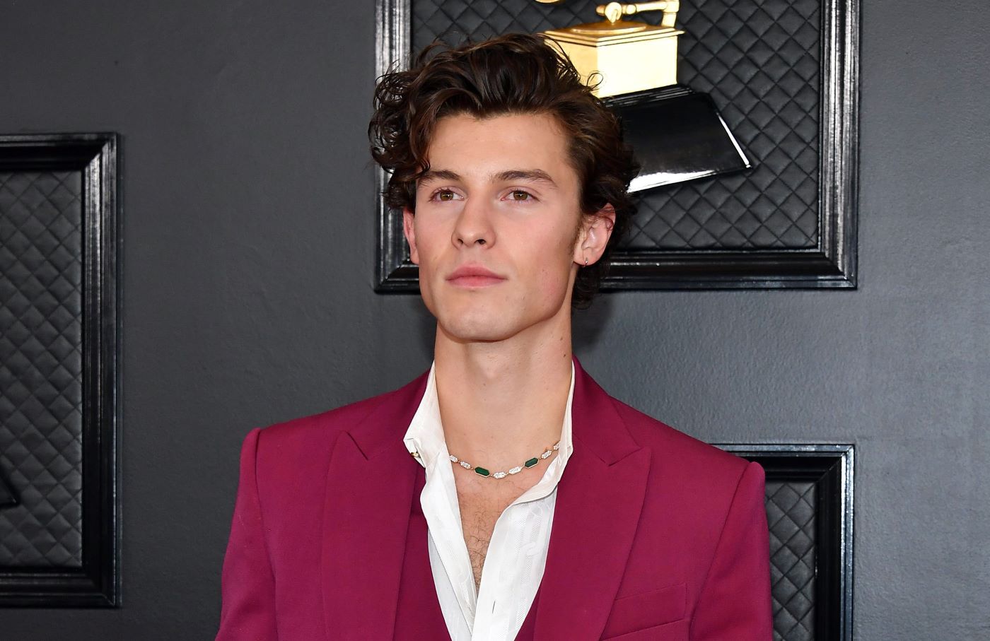 Shawn Mendes standing in front of a black background with golden awards on it wearing a red suit with a white undershirt.