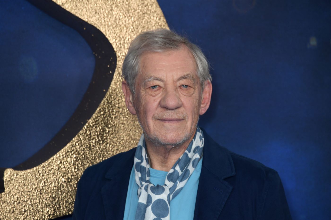 Sir Ian McKellen wearing a blue undershirt with a dark blue jacket and a white and teal ascot tie in front of a dark blue and gold background.
