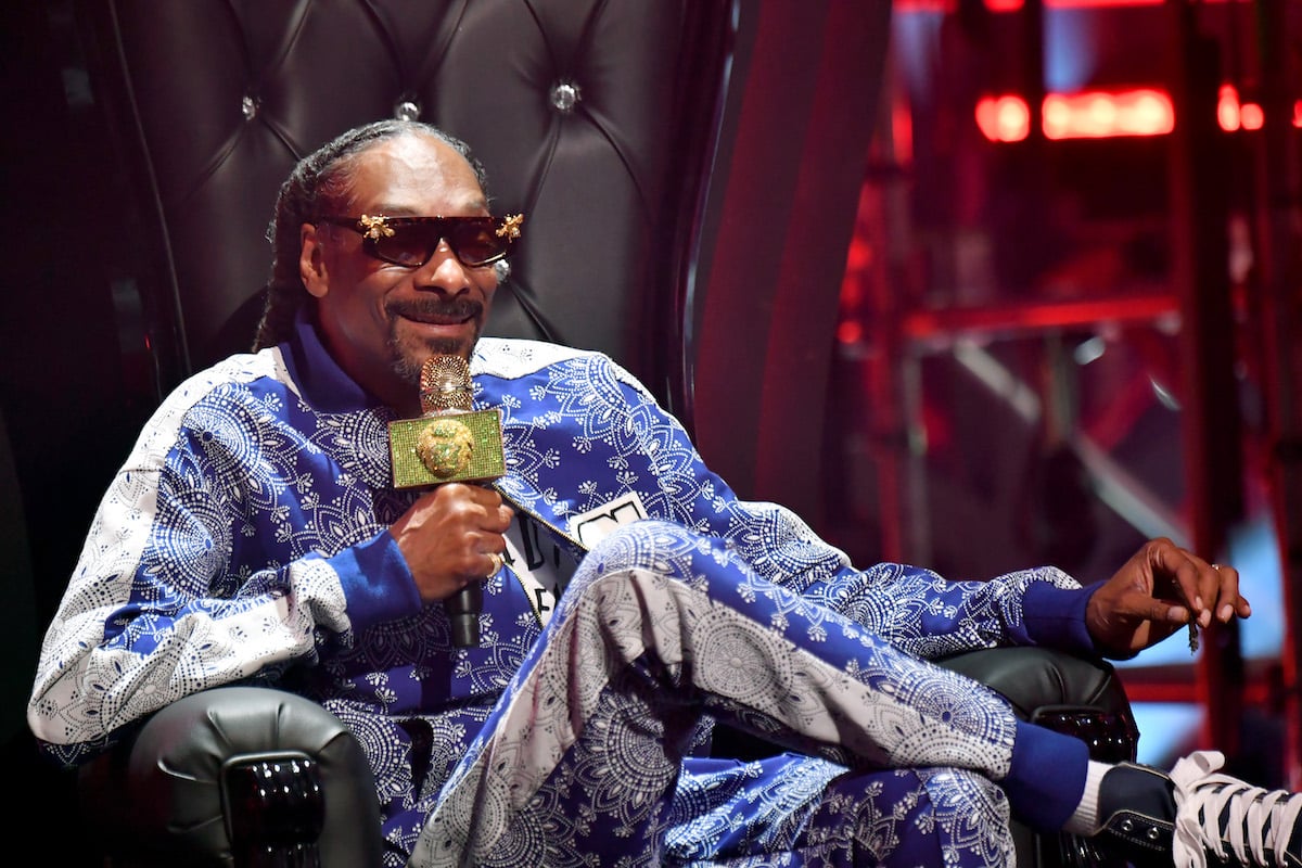 Snoop Dogg sits with his legs crossed and wearing sunglasses, smiling and talking into a microphone.