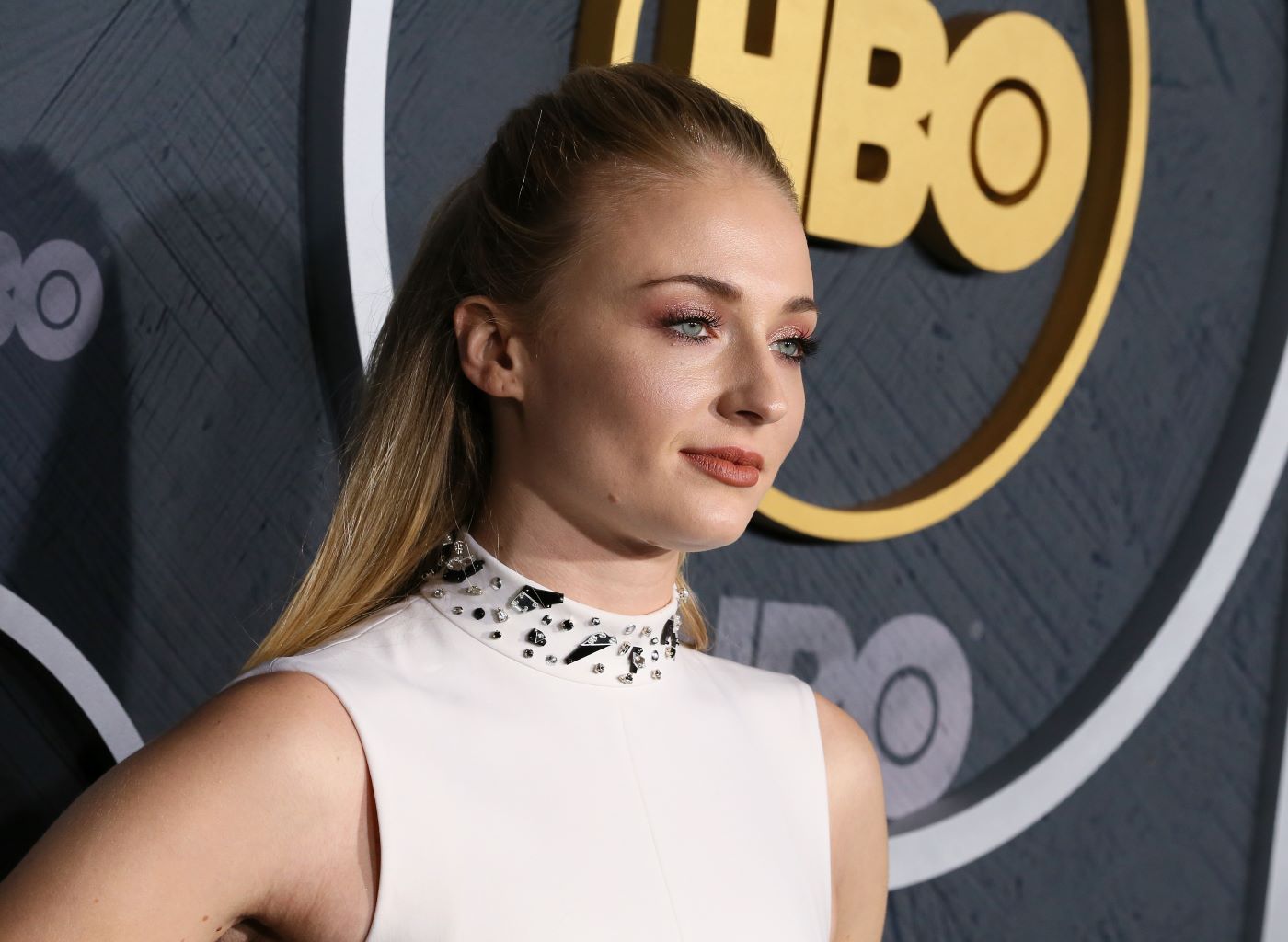 Sophie Turner is wearing a white high neck halter top dress with grey embellishments around the neck. She is standing in front of a black background with the HBO logo in gold.