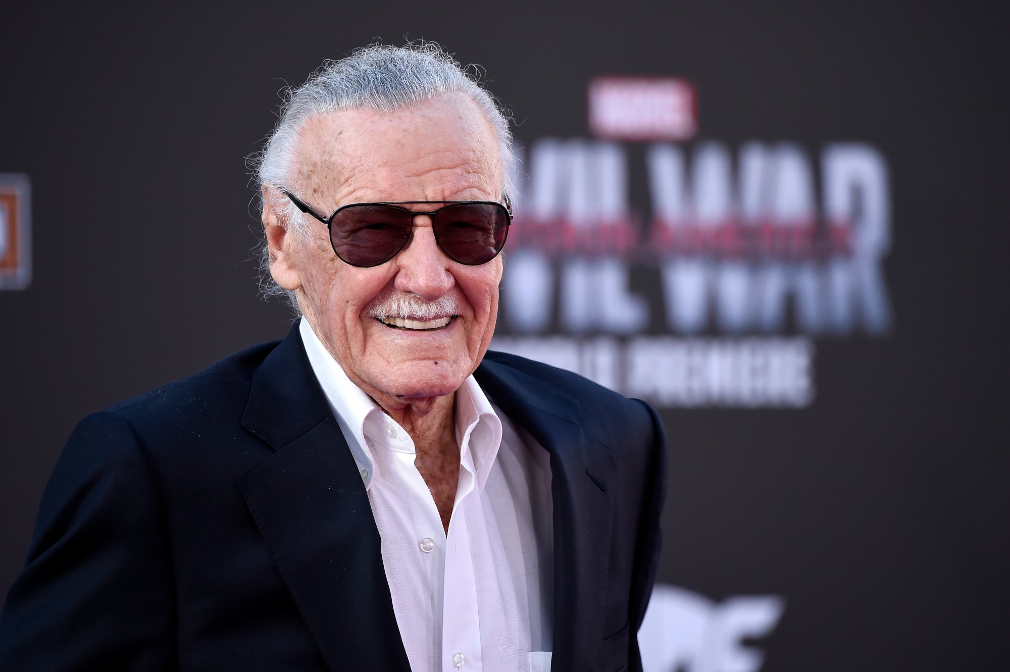 Marvel's Stan Lee wearing a black suit and smiling in front of a 'Captain America: Civil War' wall