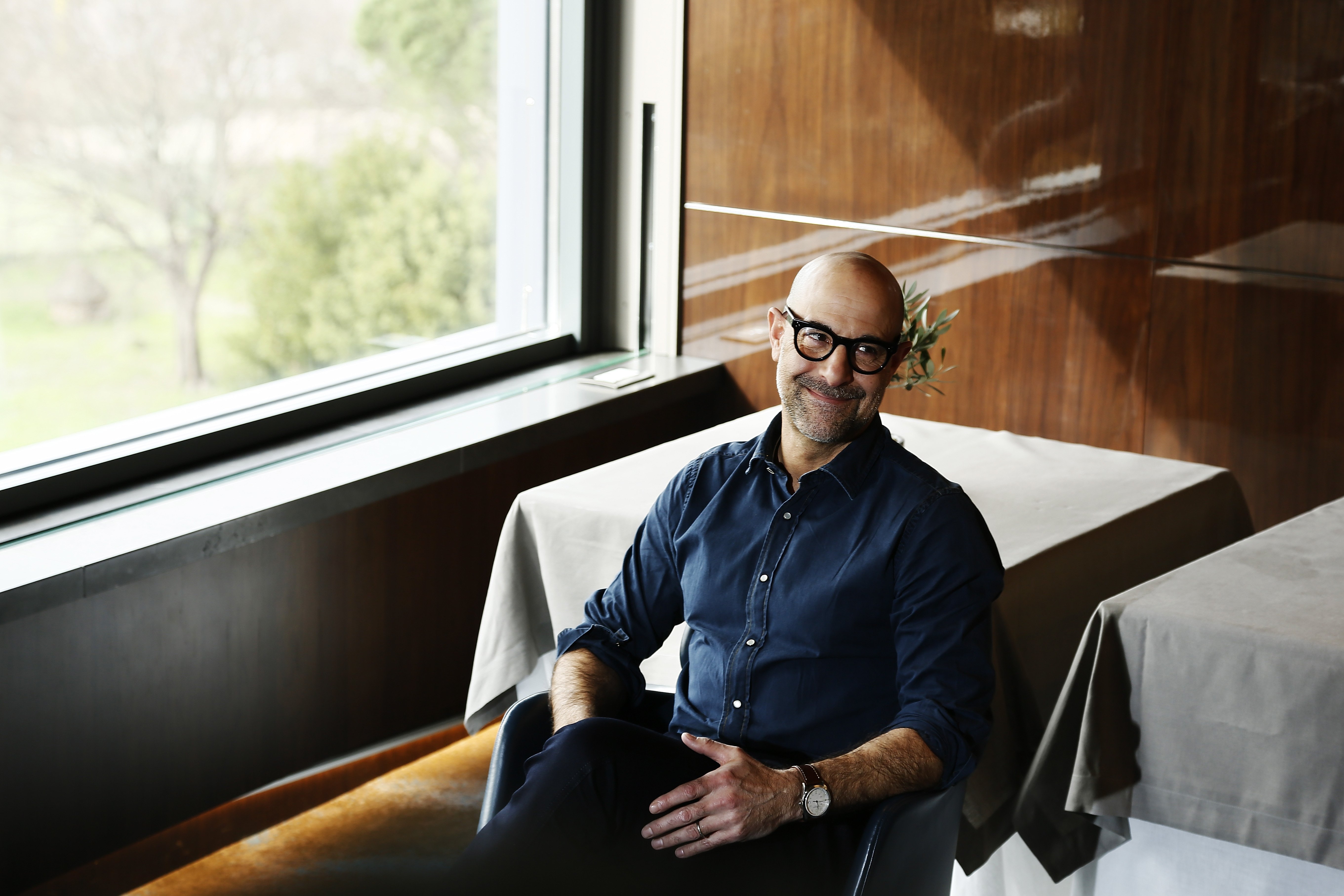 Stanley Tucci smiles sitting in a chair beside a table covered with a white tablecloth