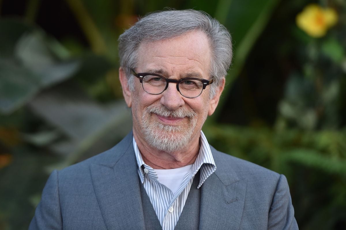 Steven Spielberg wears a gray suit and smiles in front of a leafy background