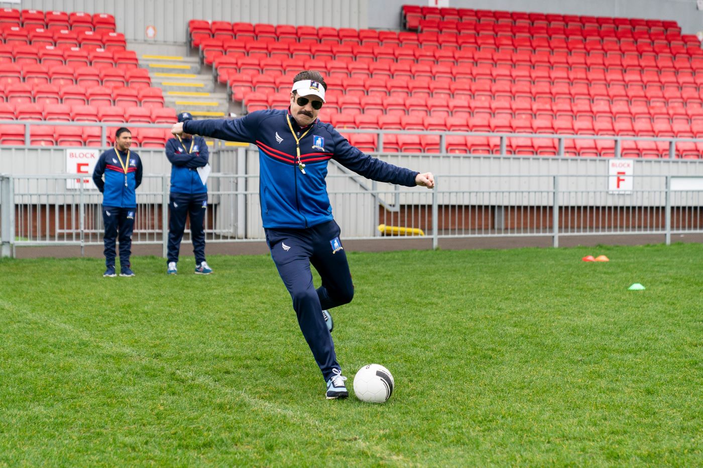 Ted Lasso as Led Tasso kicking a soccer ball on the soccer field.