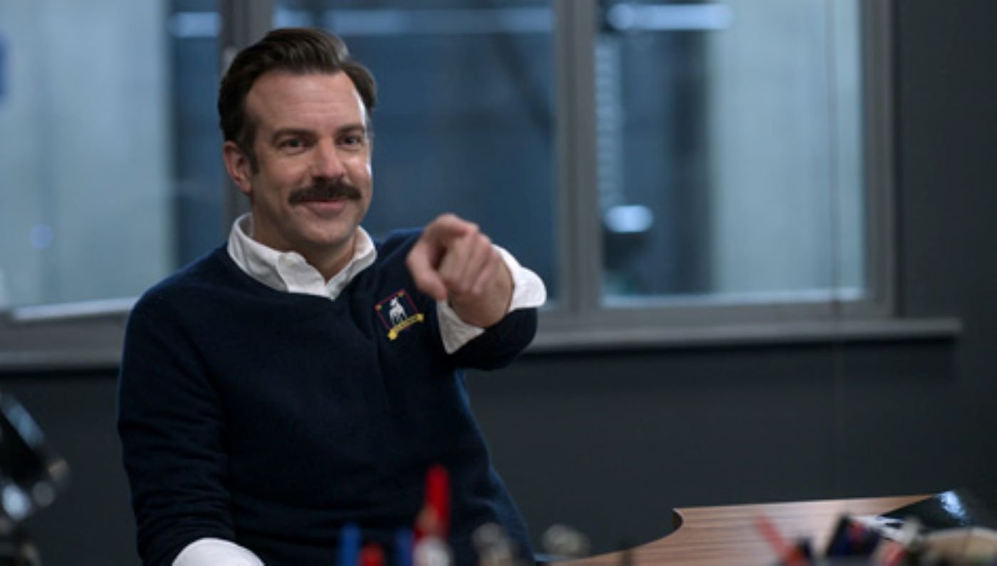 Ted Lasso sits in a blue sweater with a white dress shirt underneath in his office.