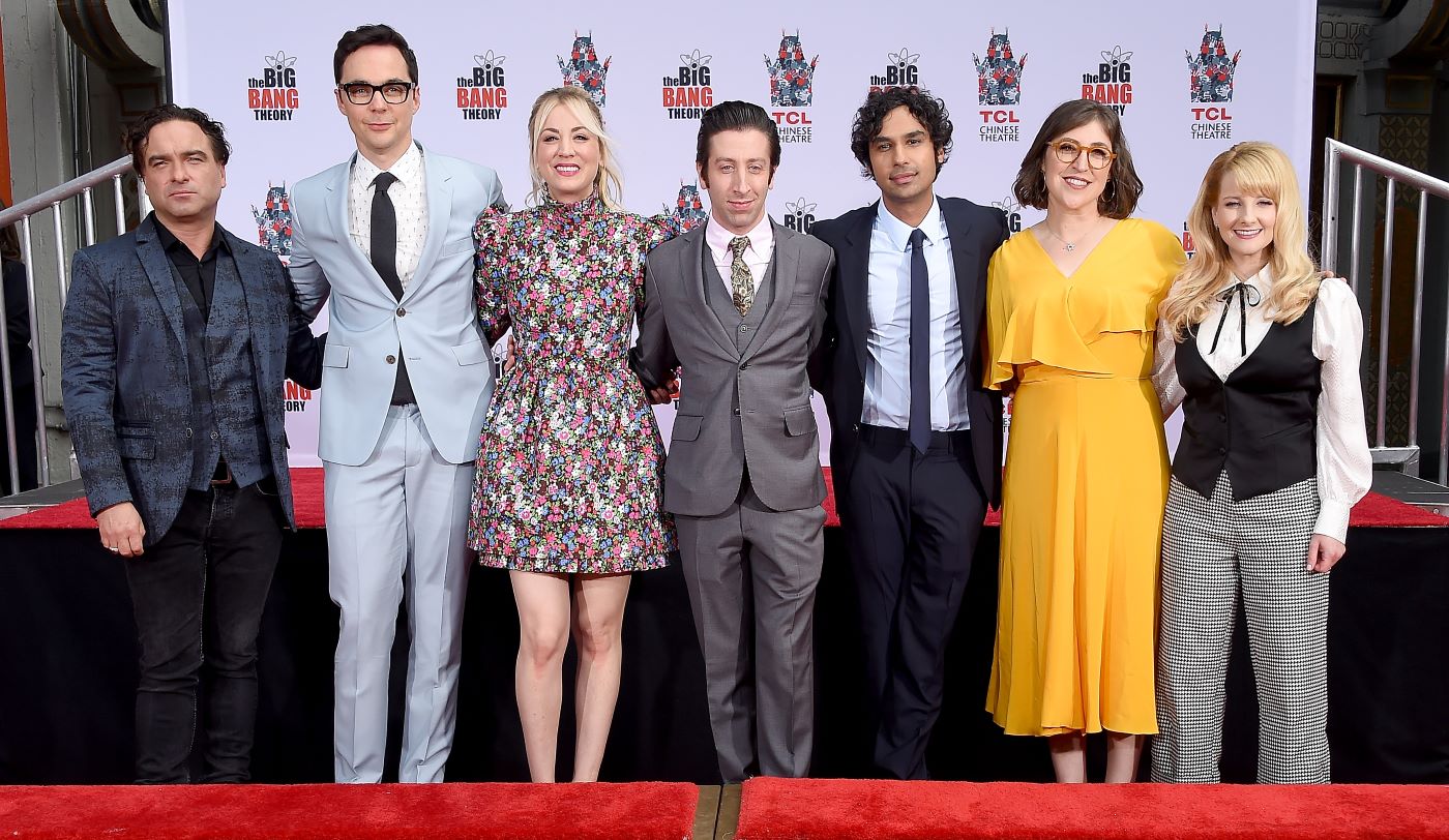 The main seven actors of 'The Big Bang Theory' standing in front of a white black and red background dressed professionally.