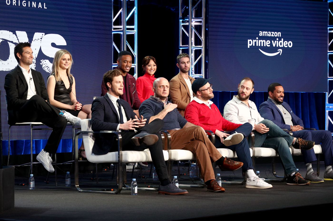 A few members of the cast of 'The Boy's' sit on a stage in white chairs against a blue background.