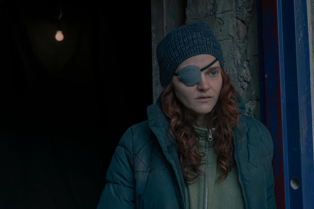 Madeline Brewer as Janine in 'The Handmaid's Tale' Season 4 Episode 5, 'Chicago.' She wears a grey zip-up hoodie with a thin, faded blue puffer jacket over it. She also wears a grey knit beanie and a grey eyepatch with a black strap as she stands in a blue and grey doorway. 'The Handmaid's Tale' Season 4 revealed Janine's backstory in episode 4, 'Milk,' which helped fans understand her more in 'The Handmaid's Tale' 'Chicago' episode.